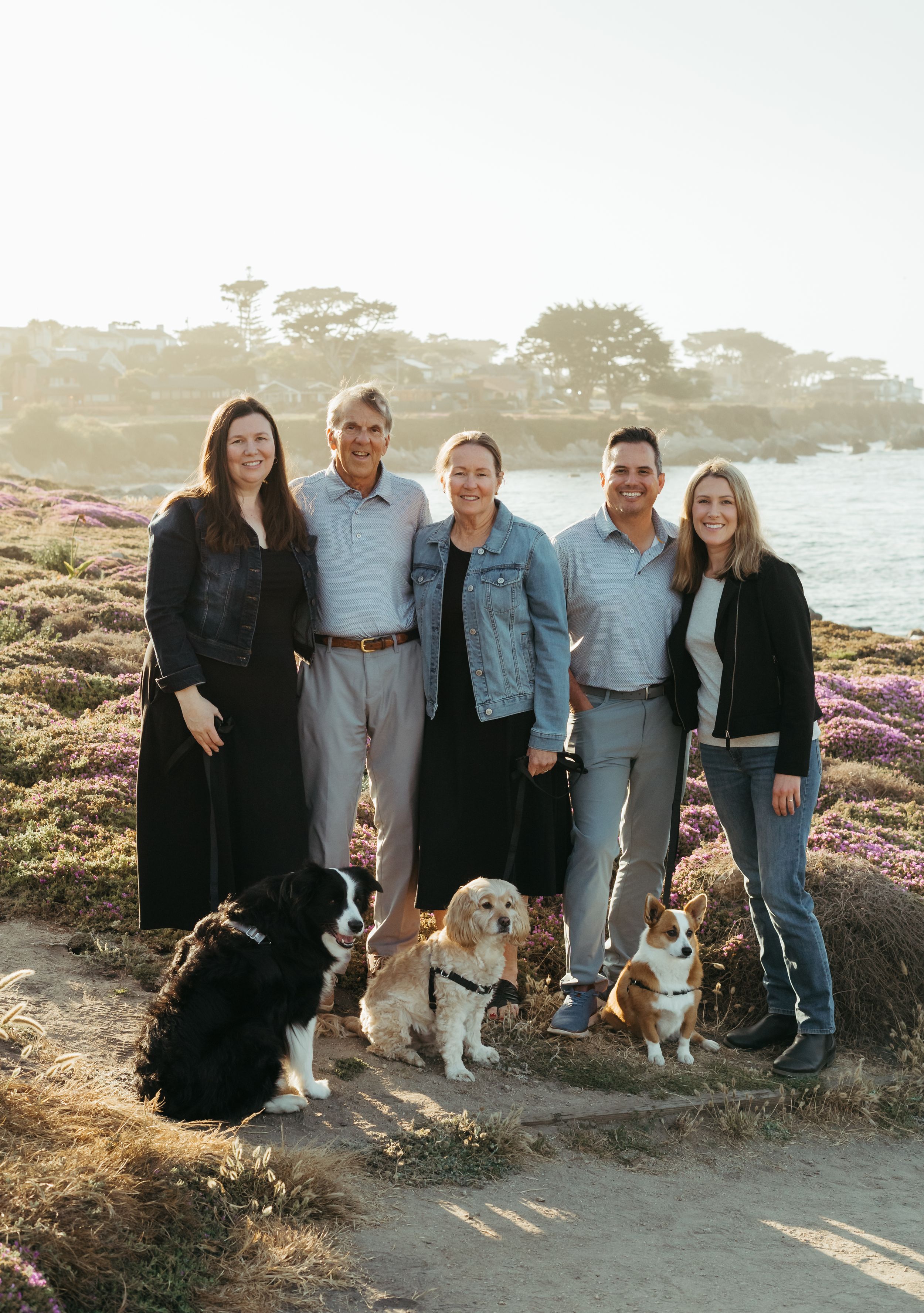 Group portrait with three dogs standing on coastal trail with ocean and cypress trees in background at sunset.