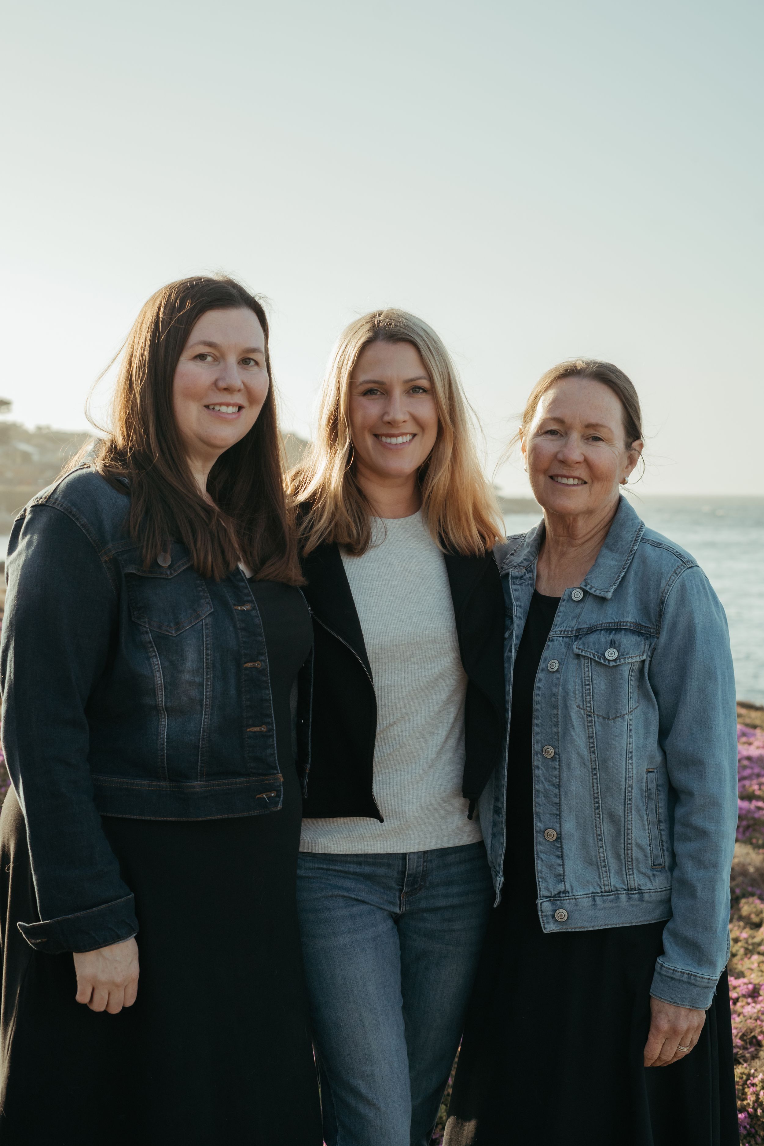 Three people in denim jackets pose together at sunset by the ocean on a rocky coastline.