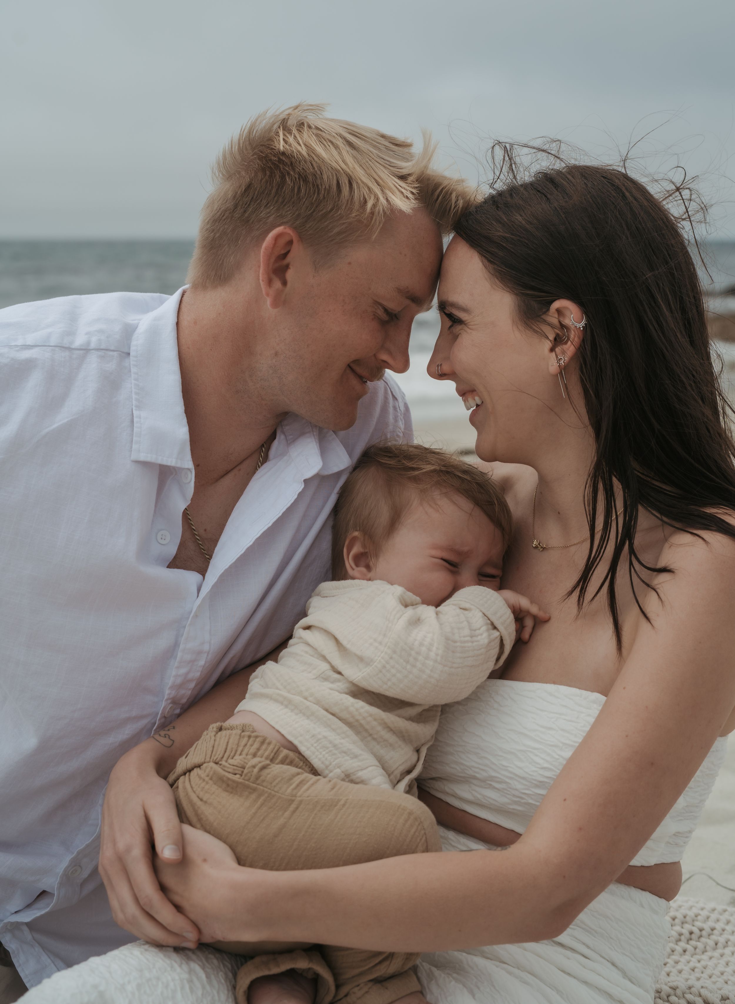 A tender family moment on the beach as parents cuddle their sleeping baby in white and beige tones.