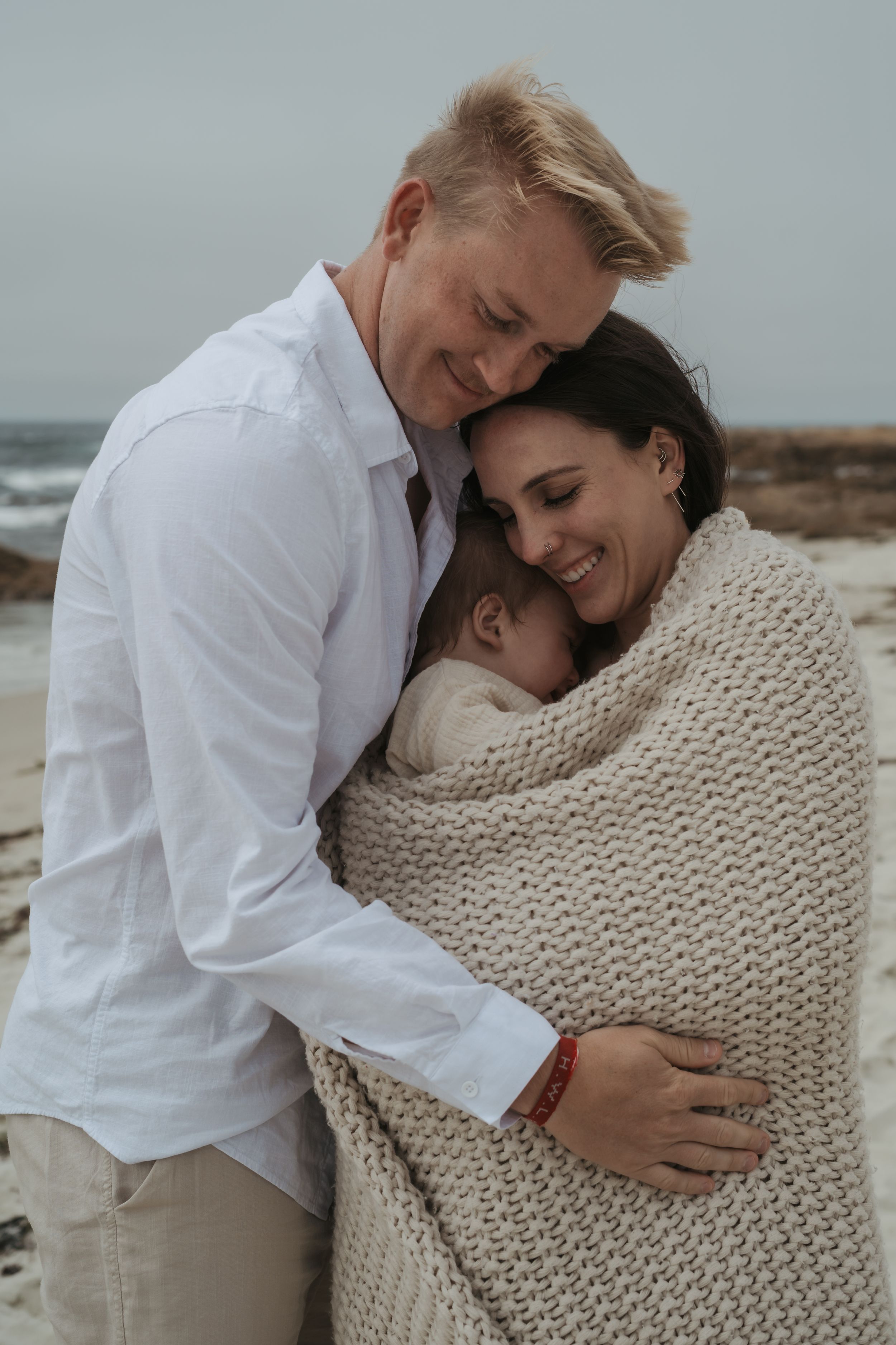 A couple embraces on the beach, with the subject wrapped in a cozy knit blanket as they share tender moments together.