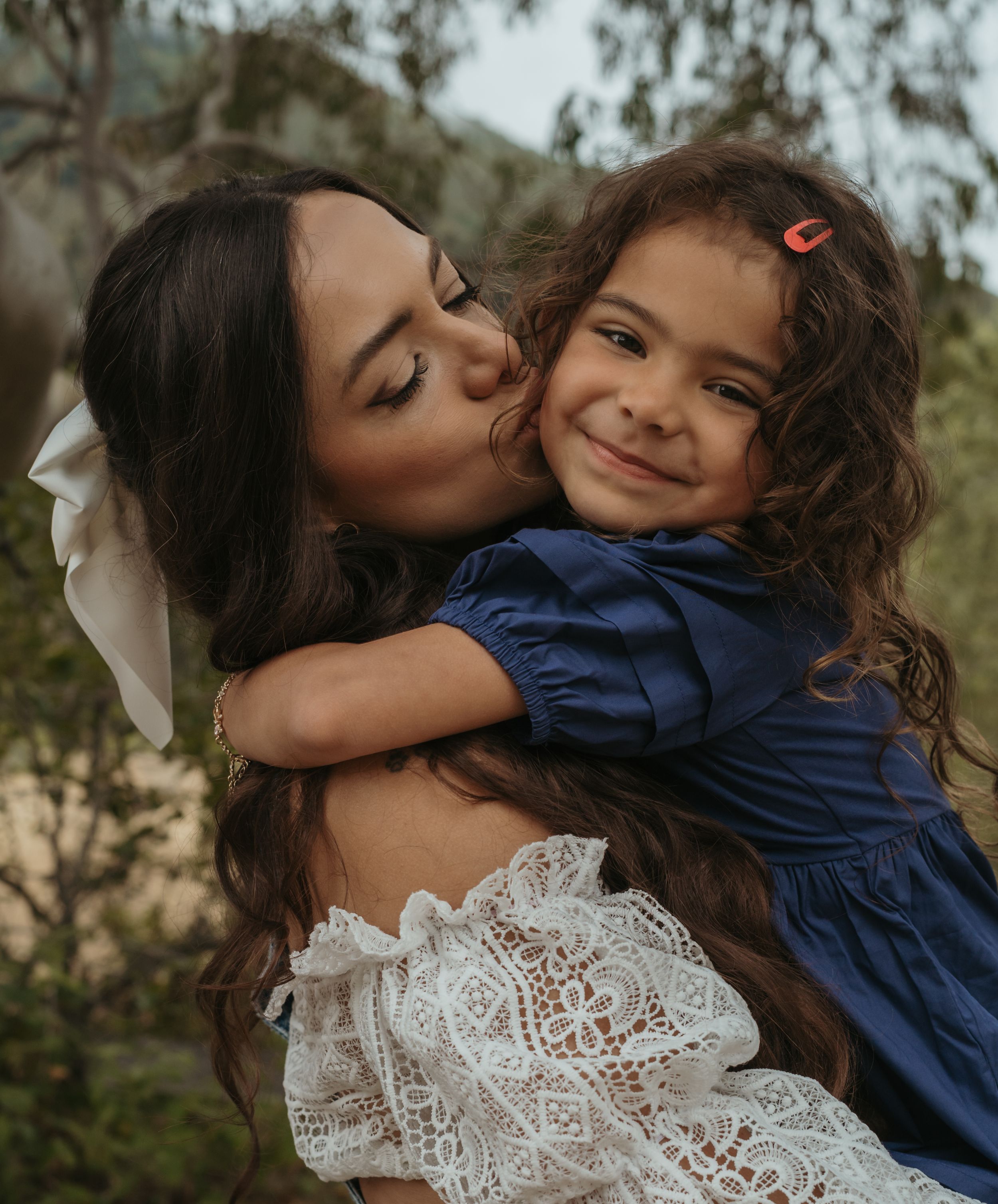 A tender family moment captured outdoors as a woman in a lace dress gives a sweet kiss to a smiling child in navy blue.