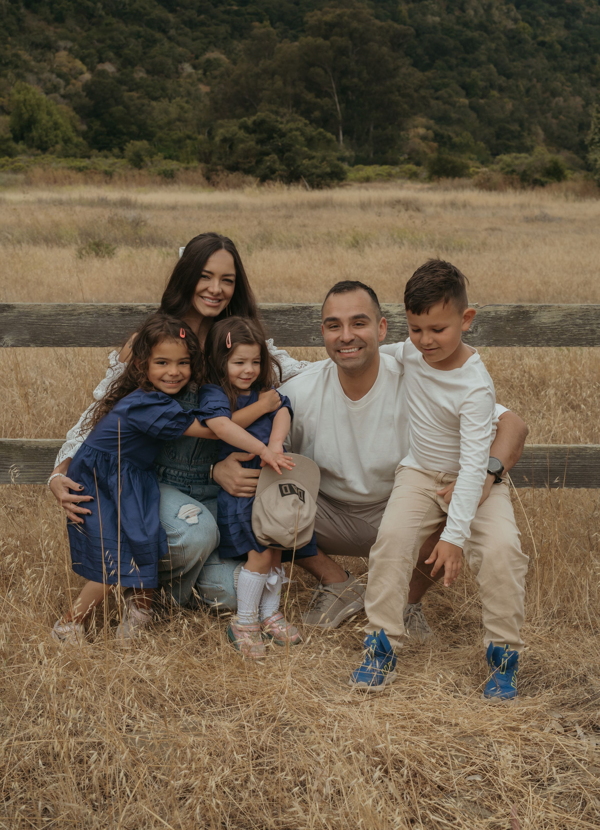 A family poses together in front of a wooden fence in a golden field with hills in the background.