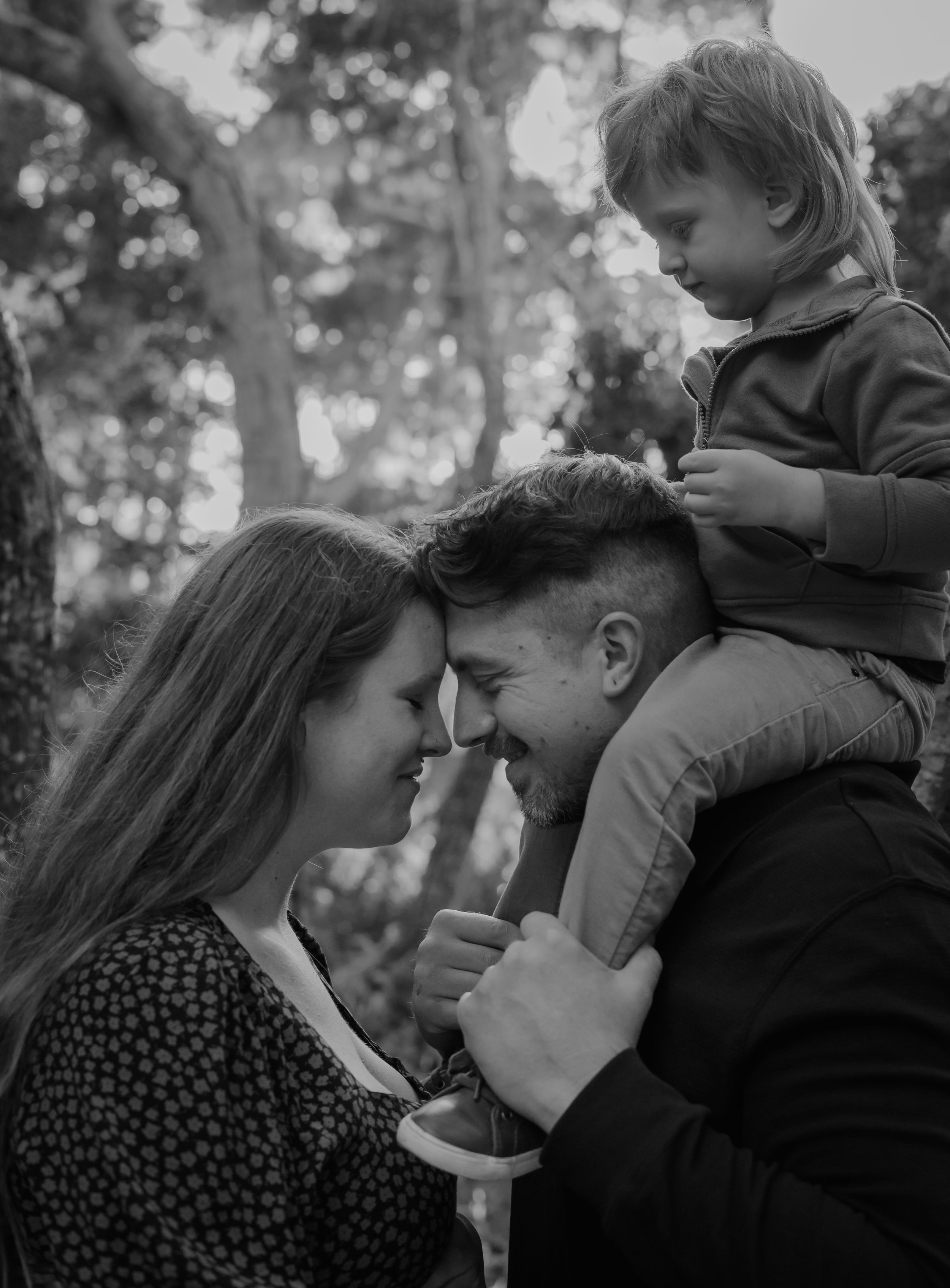 A tender black and white family portrait shows an intimate moment shared outdoors among trees.
