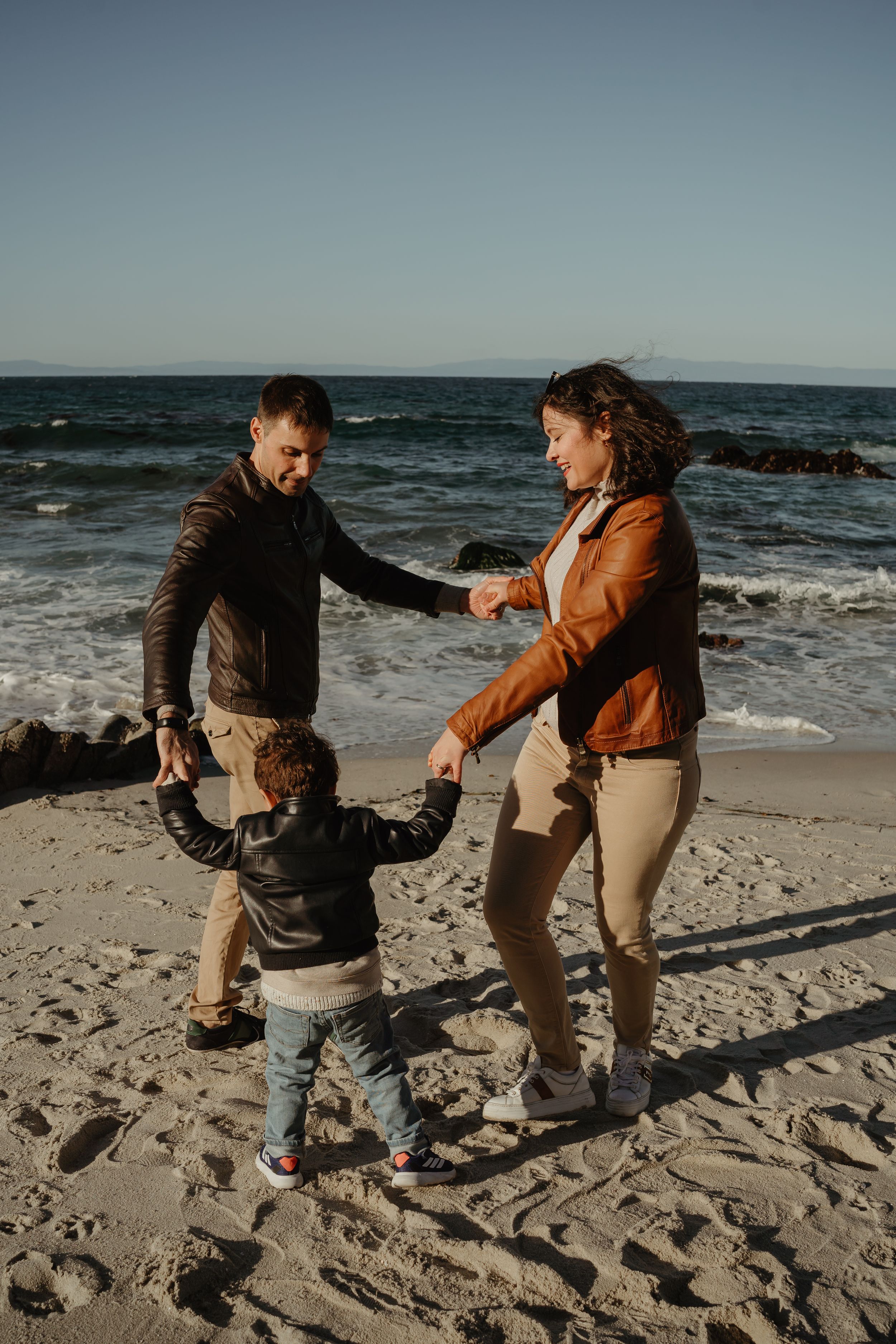 Three people holding hands and dancing in a circle on a sandy beach with ocean waves in the background.