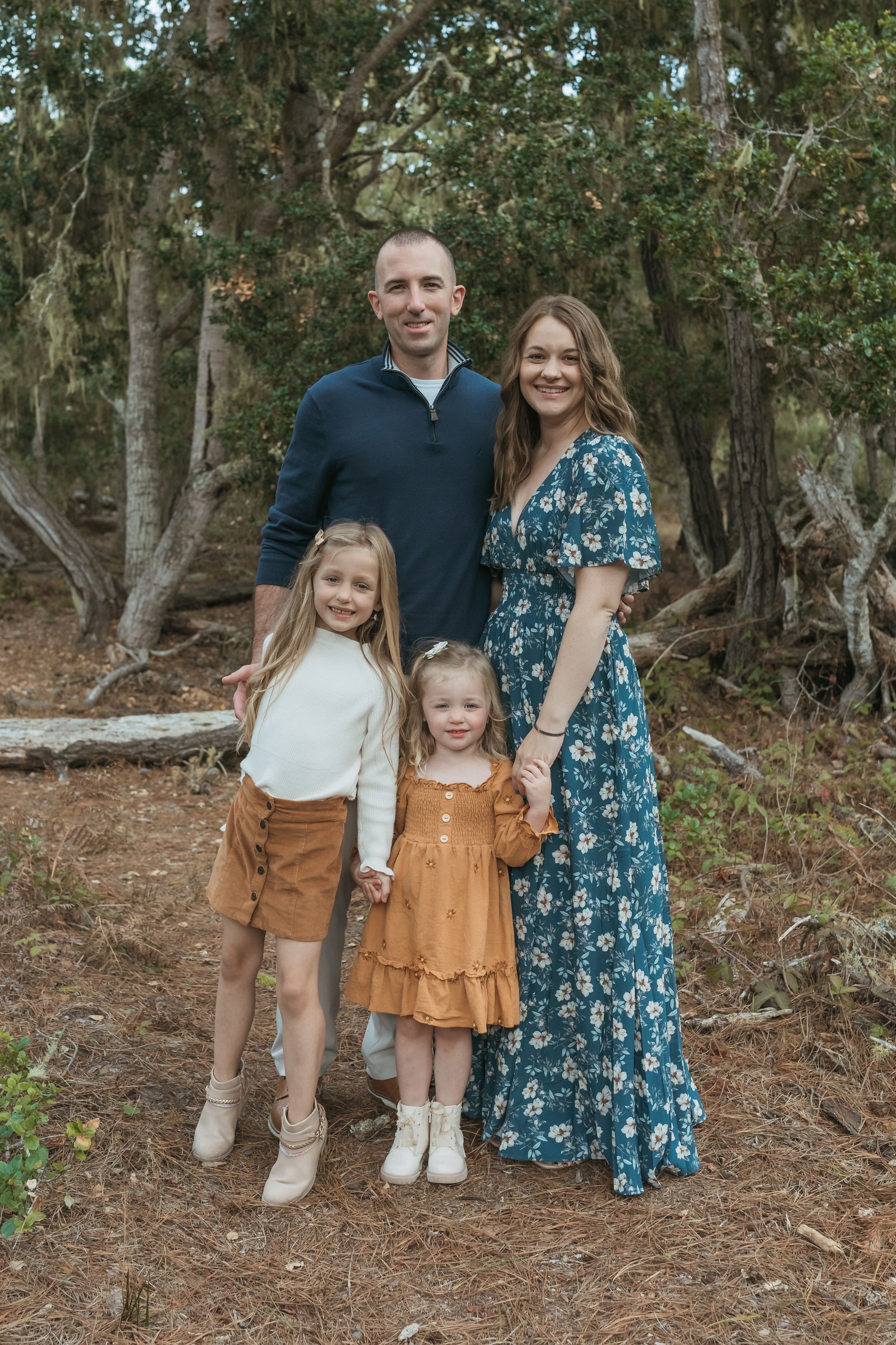 A family wearing coordinated outfits poses together in a wooded outdoor setting with eucalyptus trees in the background.