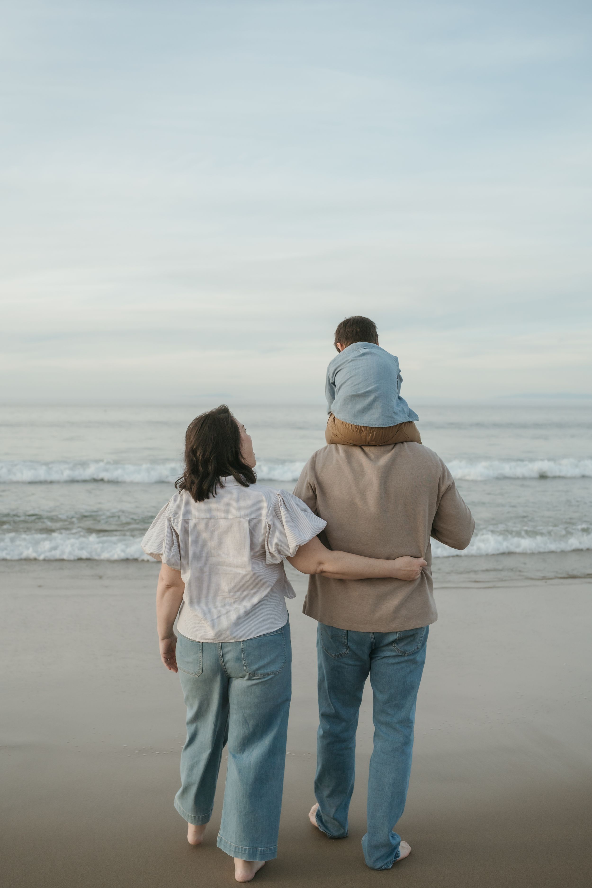 Two people walking together on a misty beach shore with their arms around each other in casual clothing.