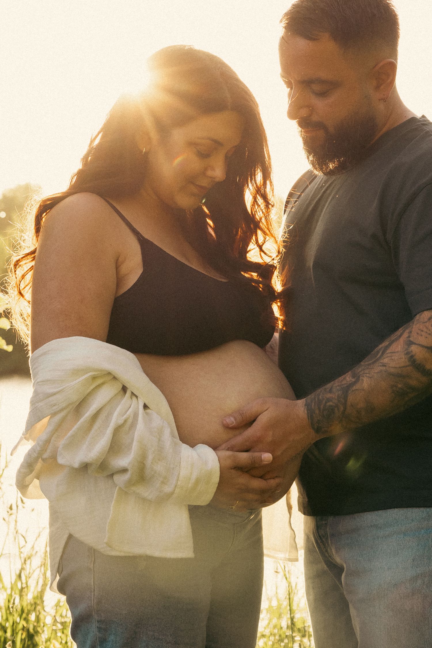 A couple shares an intimate moment during sunset as they embrace a pregnant belly in a golden-lit outdoor setting.