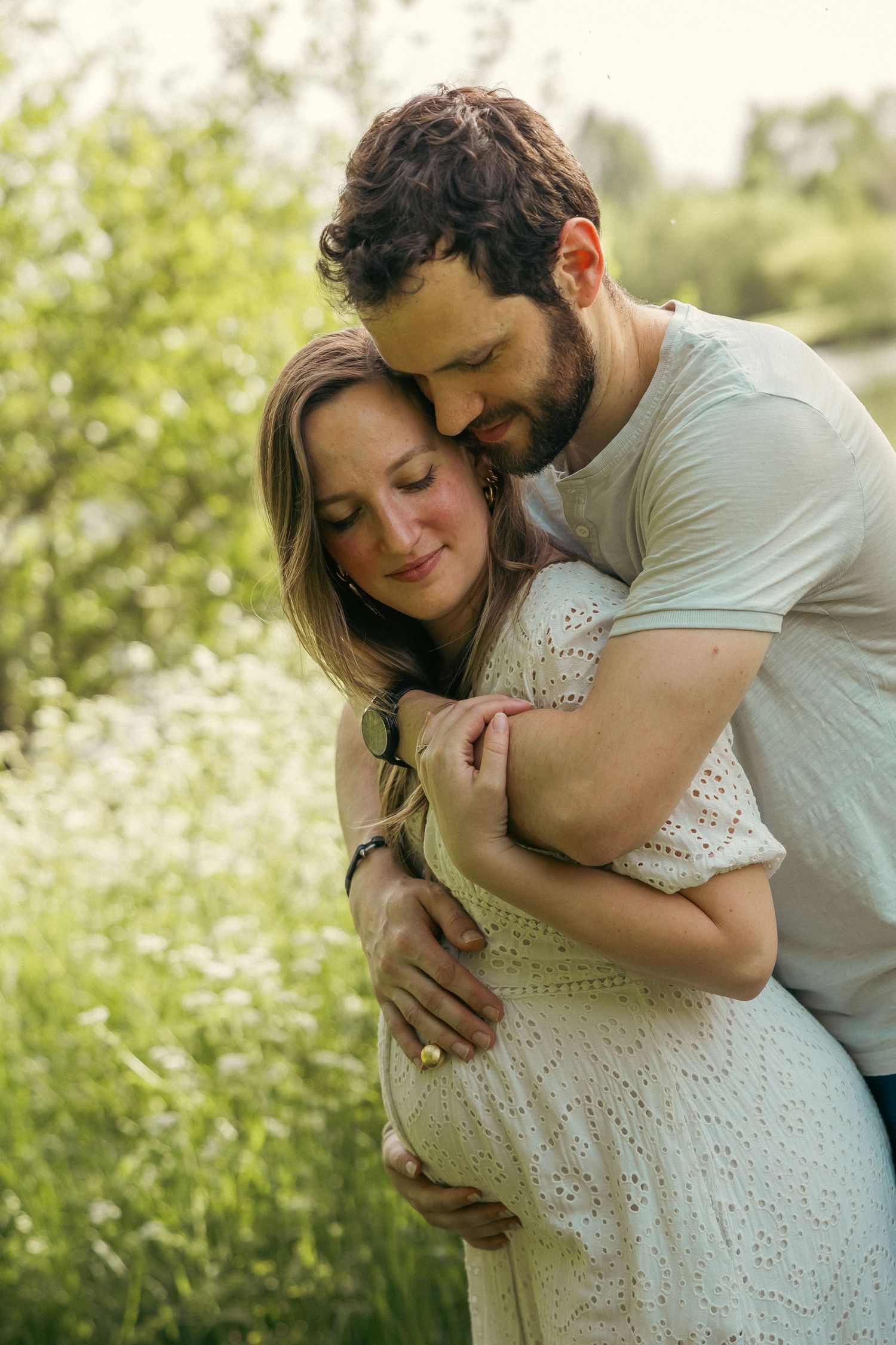 A loving couple embraces outdoors in a sunlit field, with the woman wearing a white lace dress showing her pregnancy.