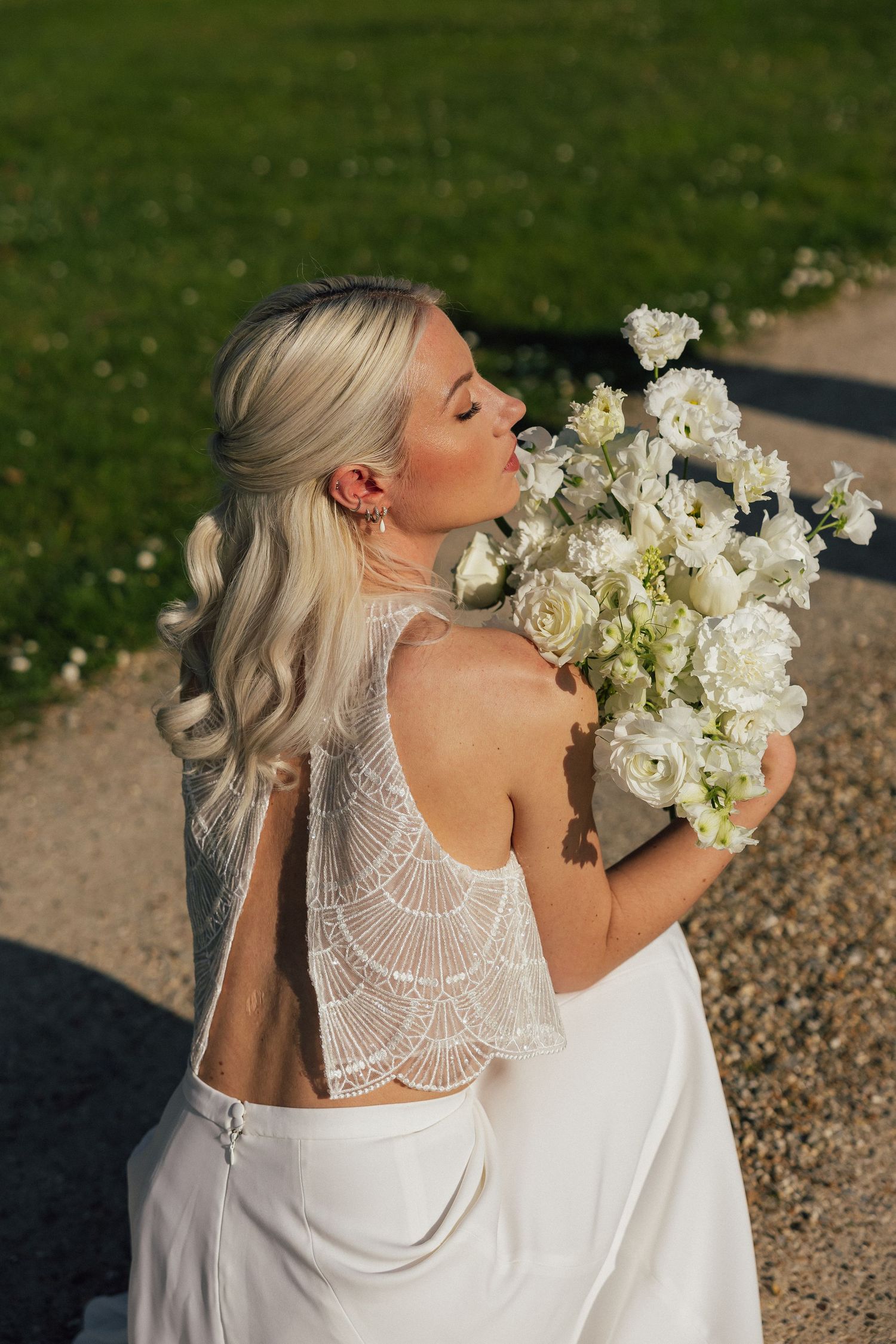 Beautiful bride in white lace gown with open back detail holds luxurious white rose and peony bouquet in golden sunlight.