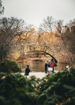 Stone bridge over water in Central Park with autumn foliage and city buildings in background.