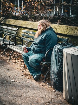 Person sitting on bench surrounded by fallen leaves and bags in urban park setting.