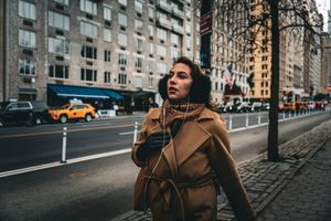 Person in brown coat walking along city street with yellow taxi and buildings in background.