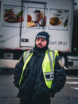 NYPD officer in reflective safety vest and winter gear stands near food truck with menu display.