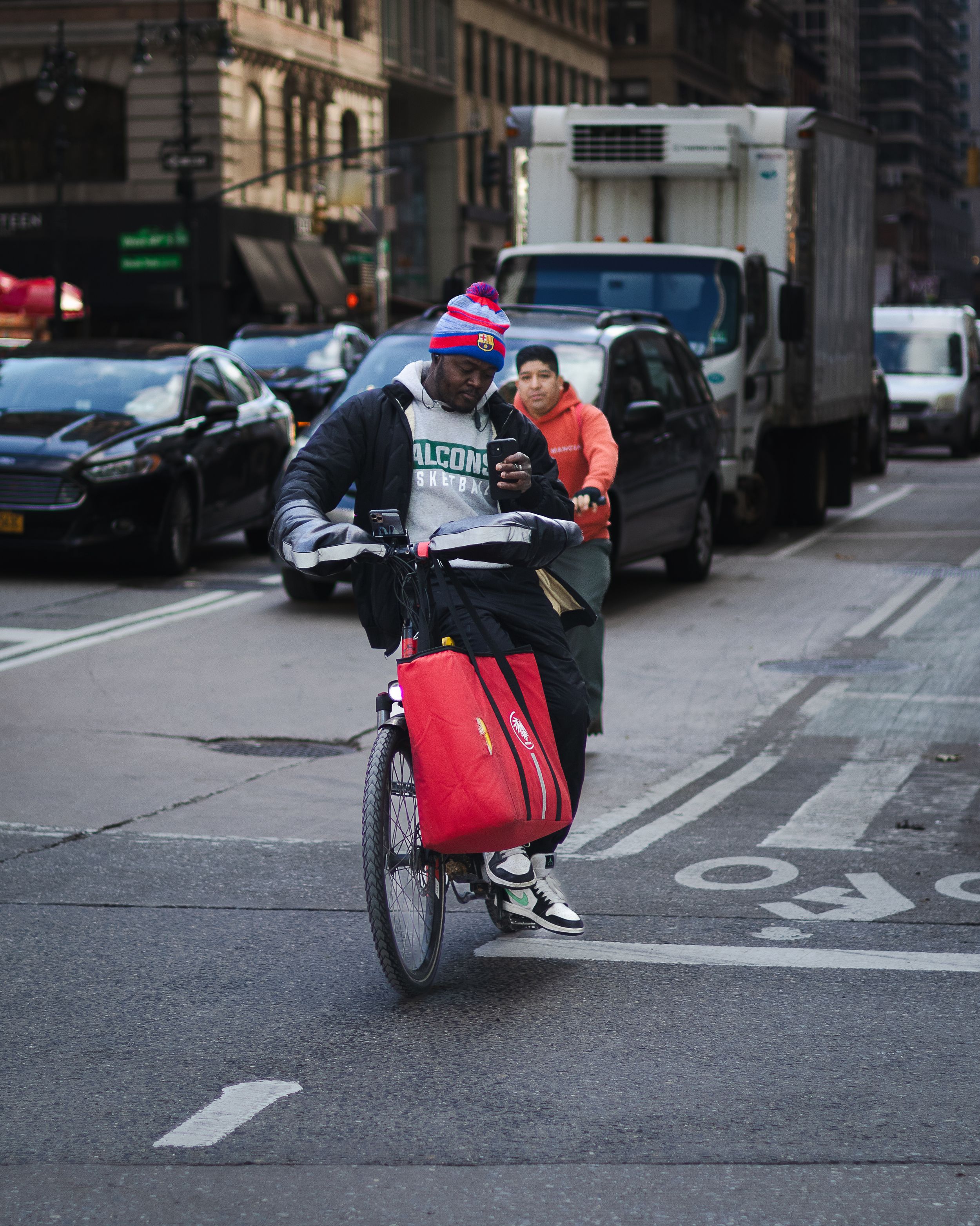 Cyclist in winter gear rides through city street with red delivery bag.