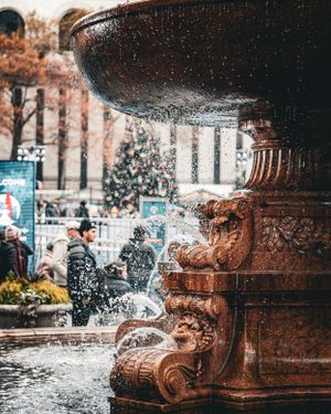 Ornate fountain sprays water in urban park setting with fall foliage.