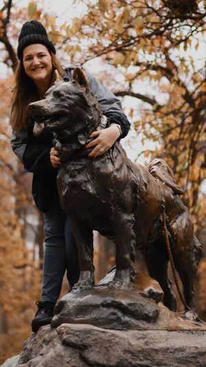 Bronze Balto statue with autumn trees and park scenery in background.