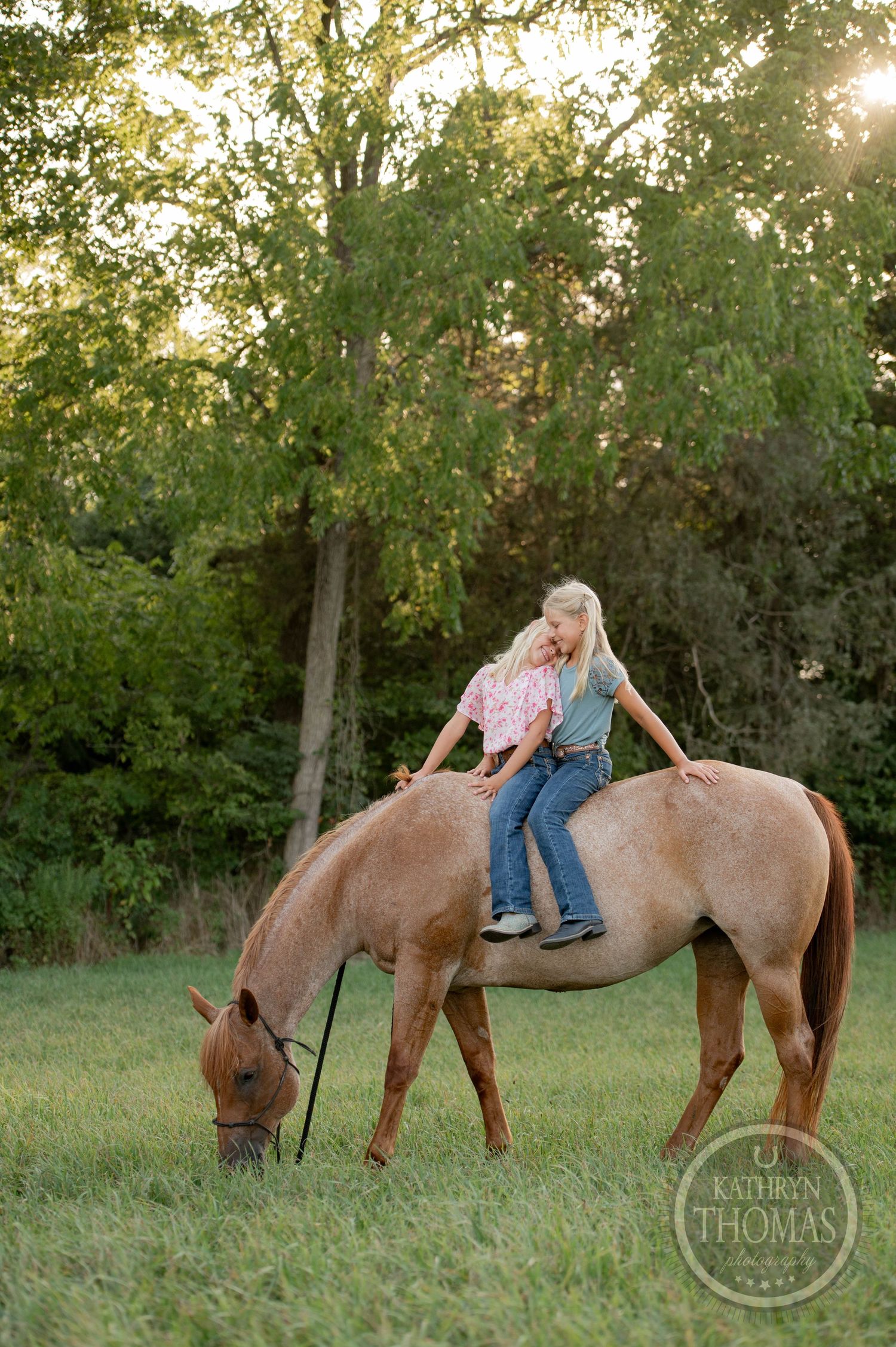 A child rides bareback on a brown horse grazing in a sunlit meadow with trees in the background.