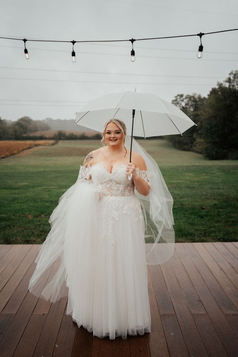 A bride in a flowing white tulle gown holds a clear umbrella while standing on a wooden deck with string lights overhead.