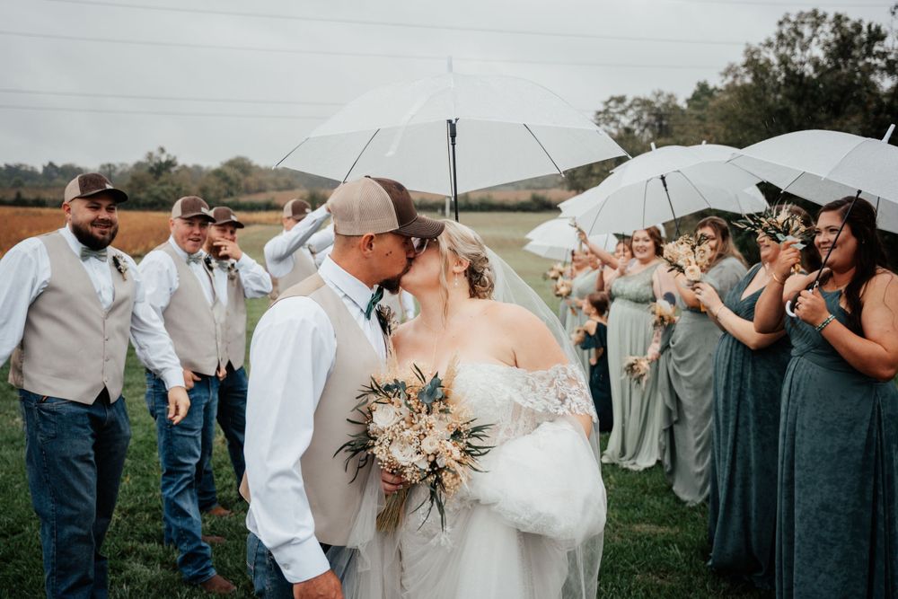 Wedding party stands in field as newlyweds share a kiss under umbrellas with bridesmaids in sage green dresses watching.