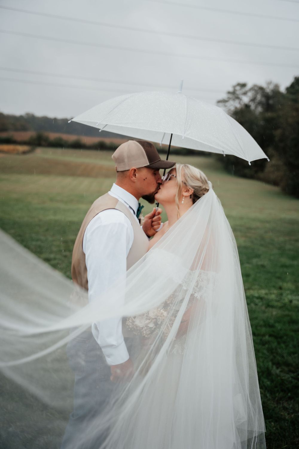 A romantic wedding moment shared under a white umbrella with a flowing veil in a countryside setting.