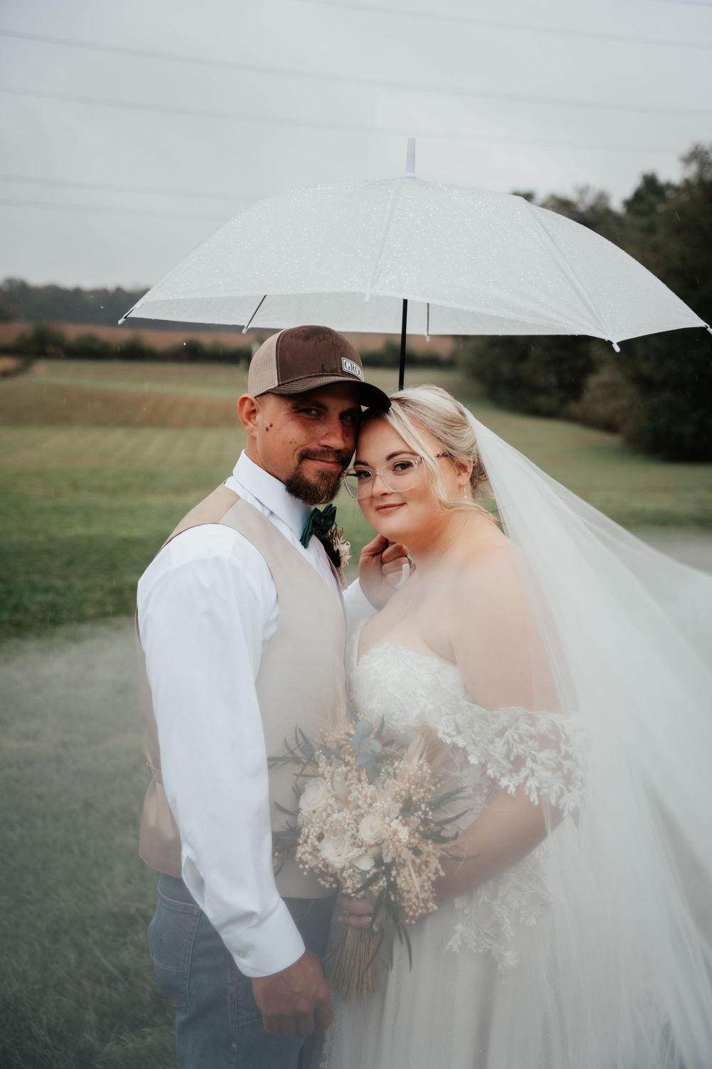 A bride and groom pose together under a white umbrella on their wedding day in a rustic outdoor setting.