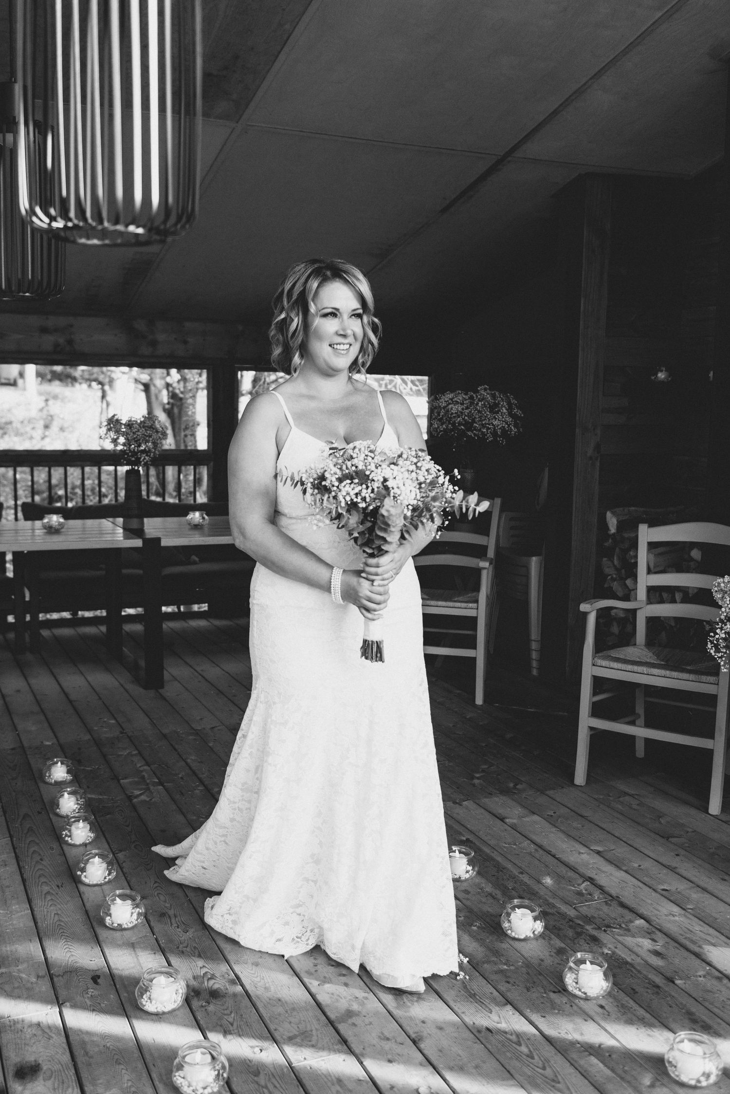 A bride in a white lace wedding dress stands on a wooden deck decorated with votives holding a flower bouquet.