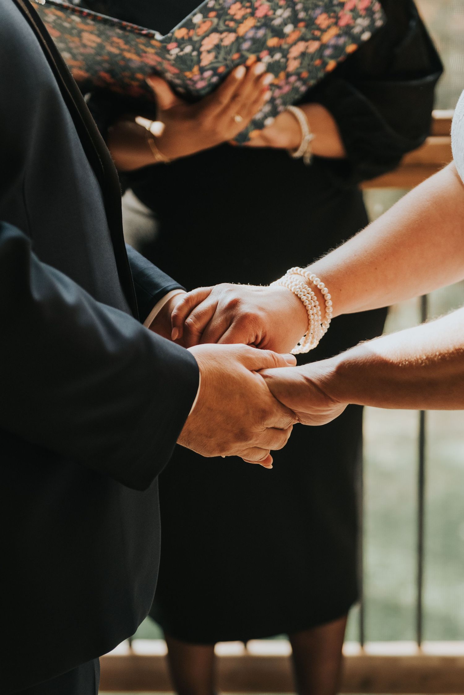 Close-up of hands joining in marriage ceremony with pearl bracelet and black formal attire visible.