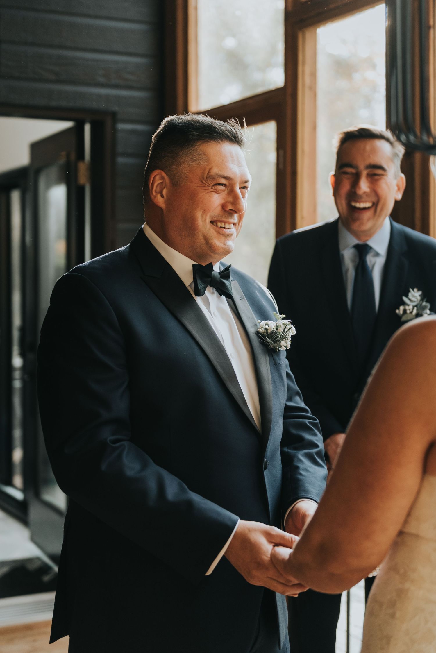 A groom in a black tuxedo with bow tie and boutonniere shares a joyful moment during a wedding celebration.
