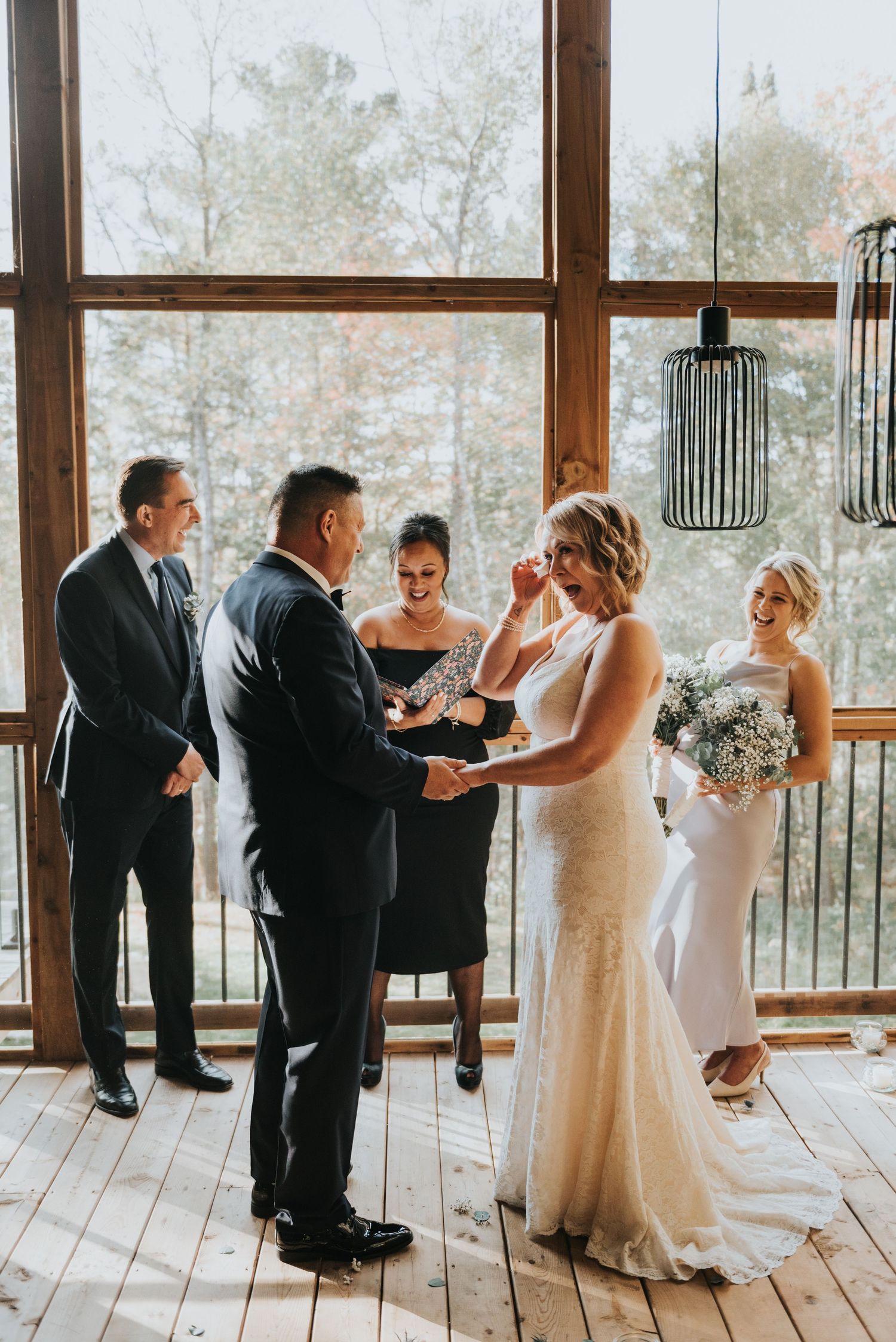 Wedding ceremony taking place in a rustic venue with large windows and pendant lights.