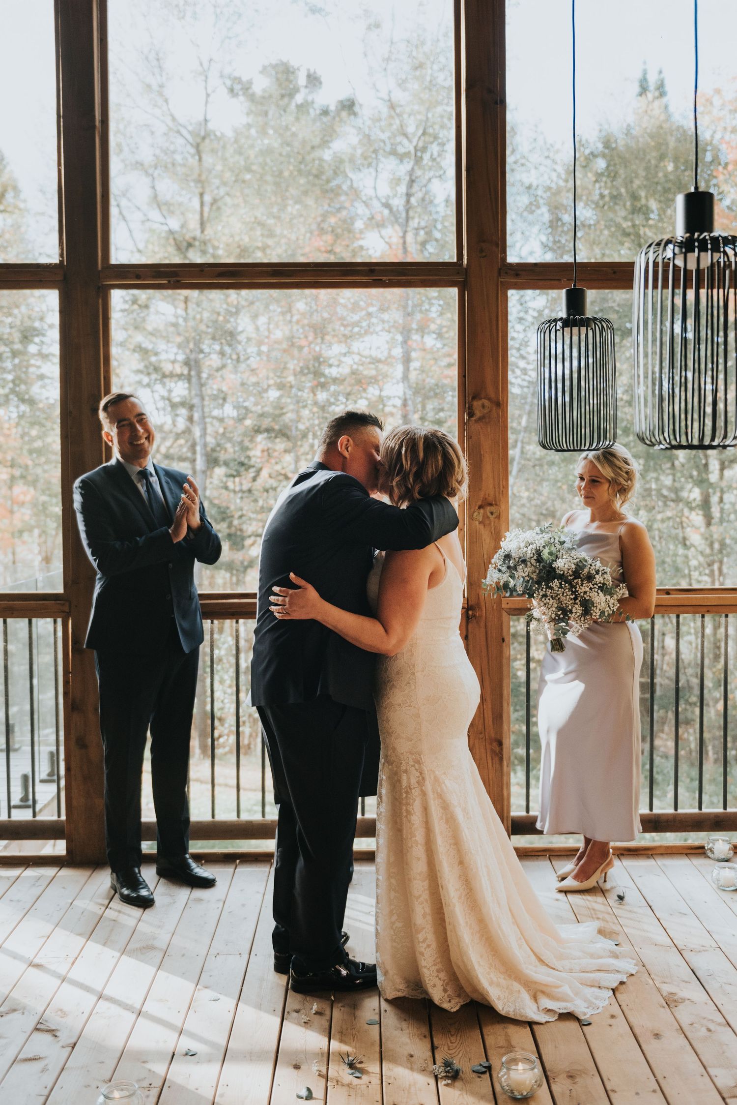 A wedding ceremony takes place in a rustic wooden venue with large windows overlooking trees in natural lighting.