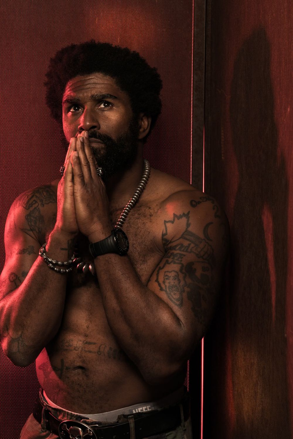 Artistic portrait of shirtless man with tattoos praying against red wall in dramatic lighting.