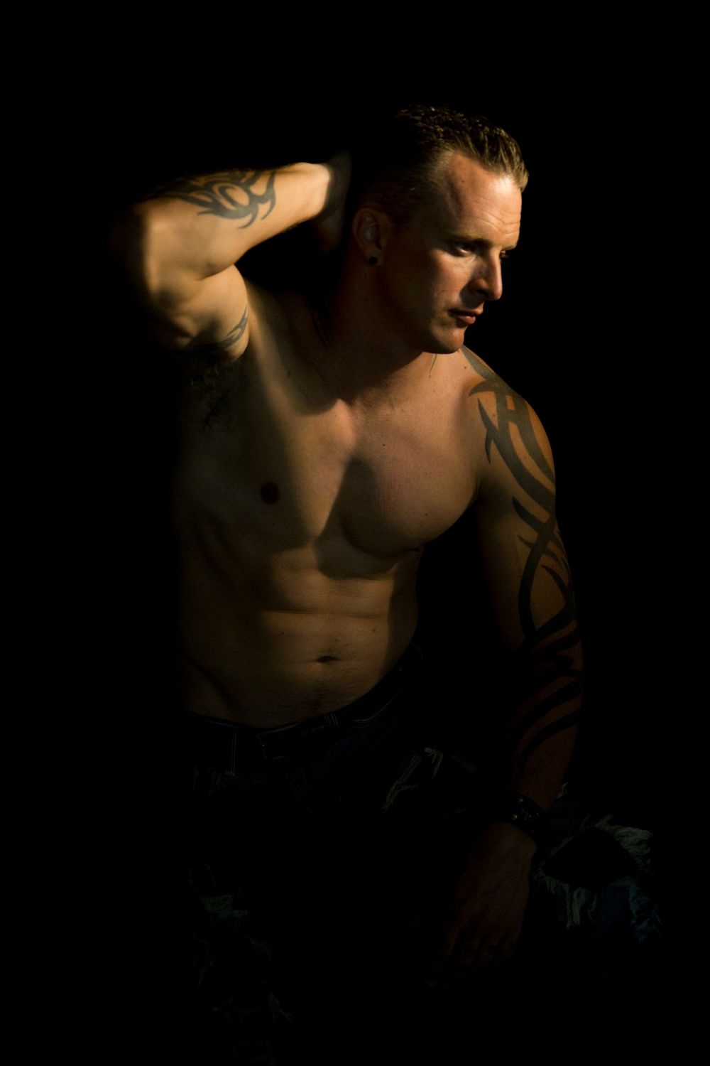 Dramatic low-key portrait of a shirtless muscular man with tattoos against a dark background.