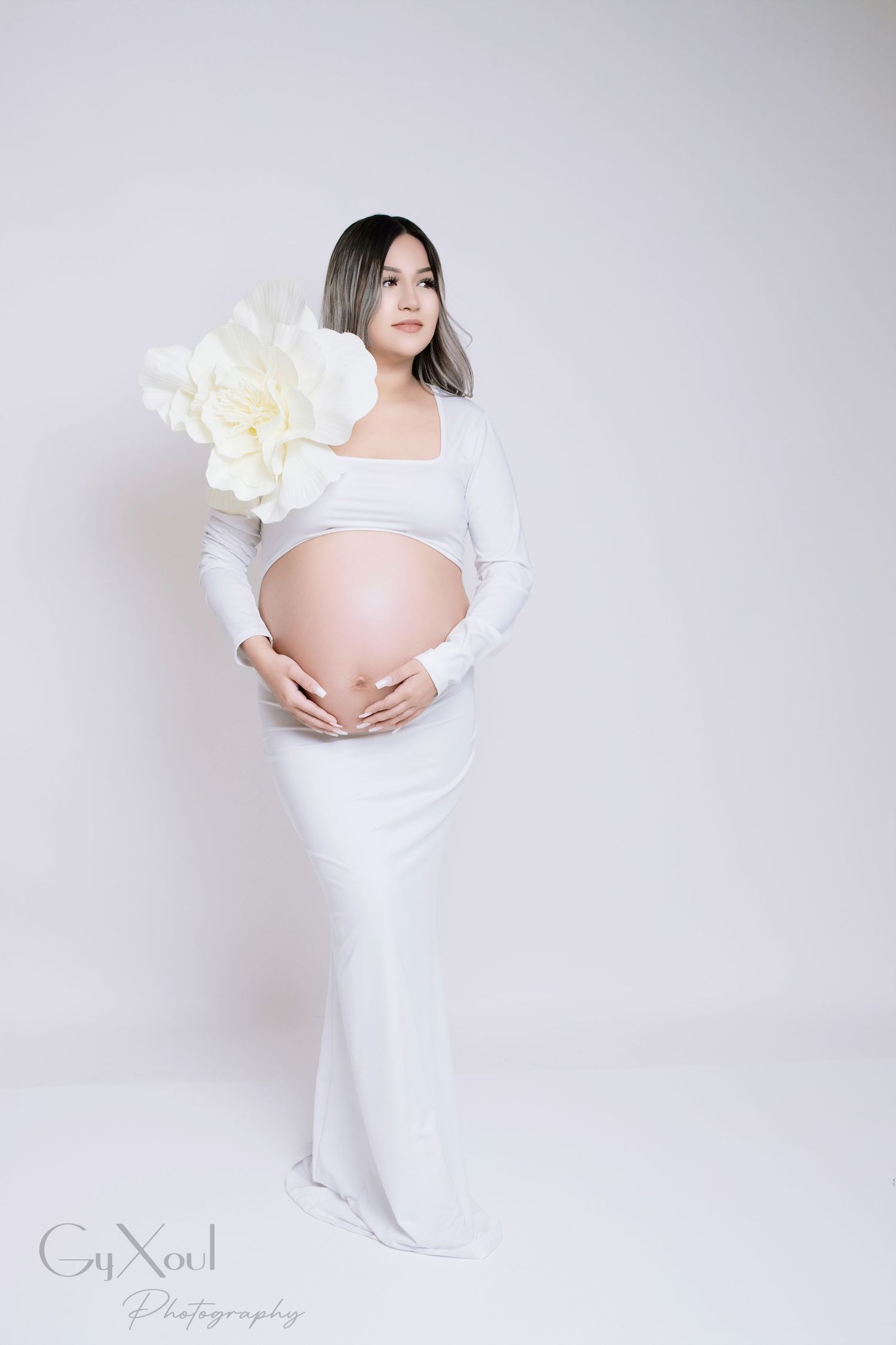 Elegant maternity portrait in white flowing dress with large floral decoration.
