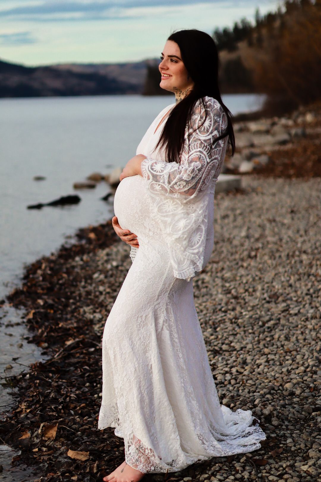 A white lace maternity gown worn on a rocky beach shoreline during sunset with mountains in the background.