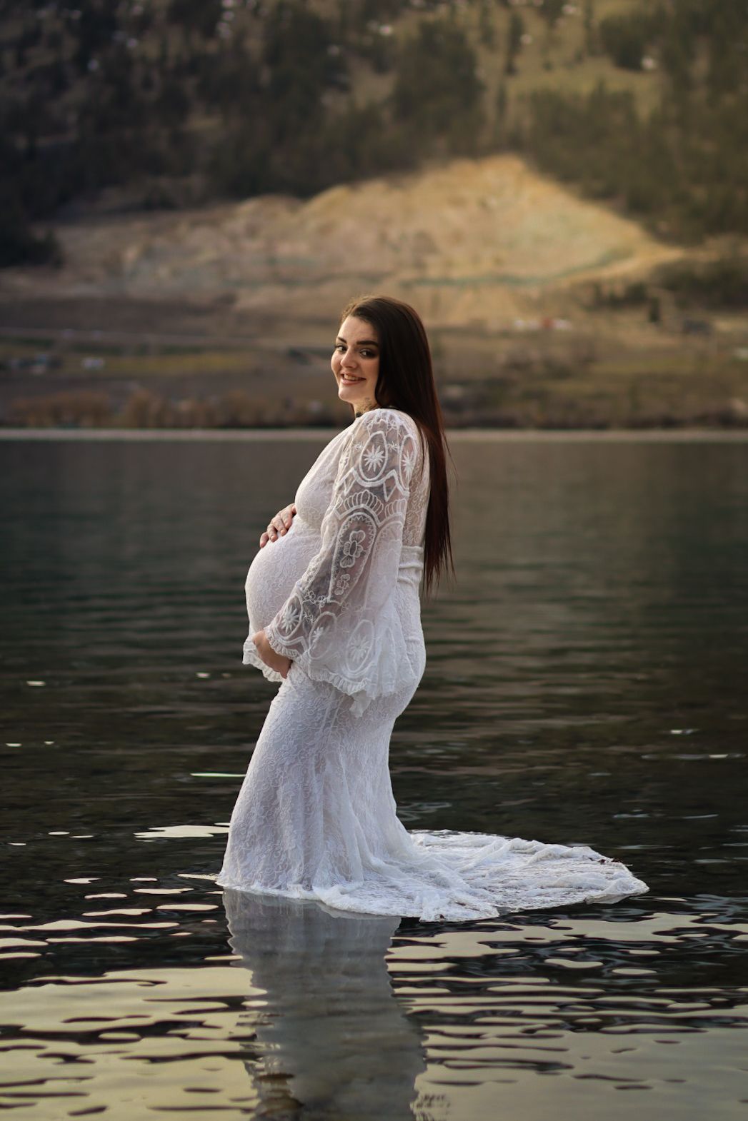 A pregnant woman in a flowing white lace dress stands in calm lake water against a scenic mountain backdrop.