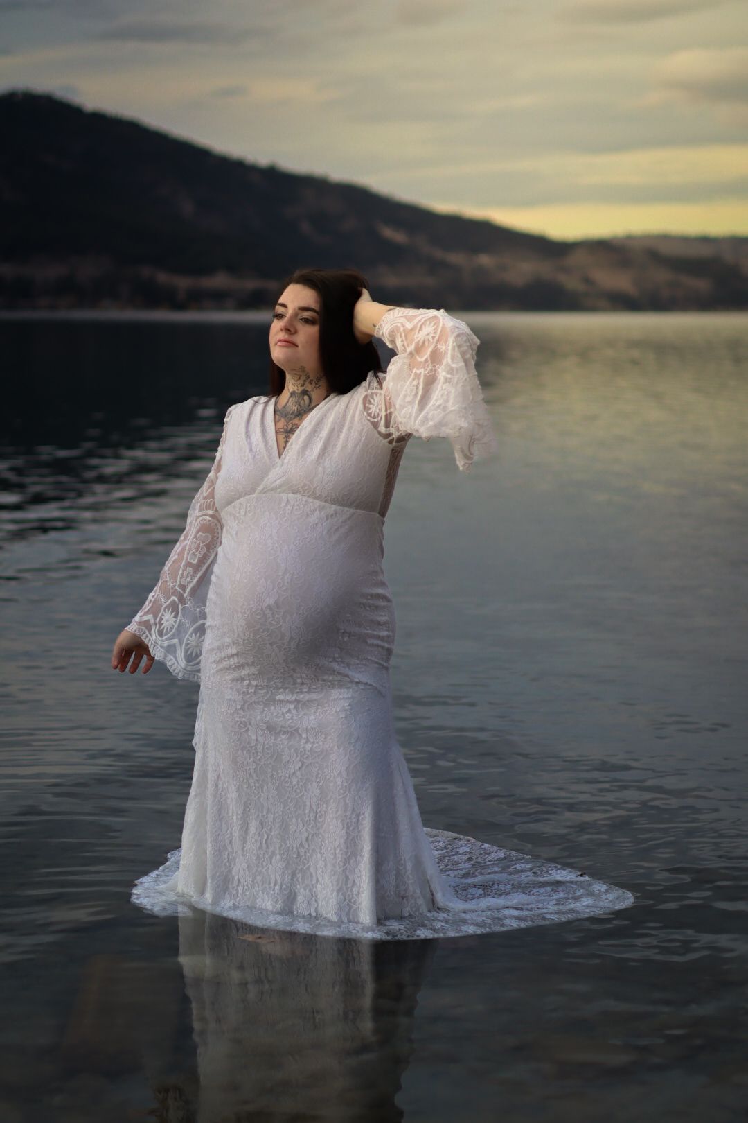 Maternity portrait of woman in elegant white dress standing in lake water against mountain backdrop.