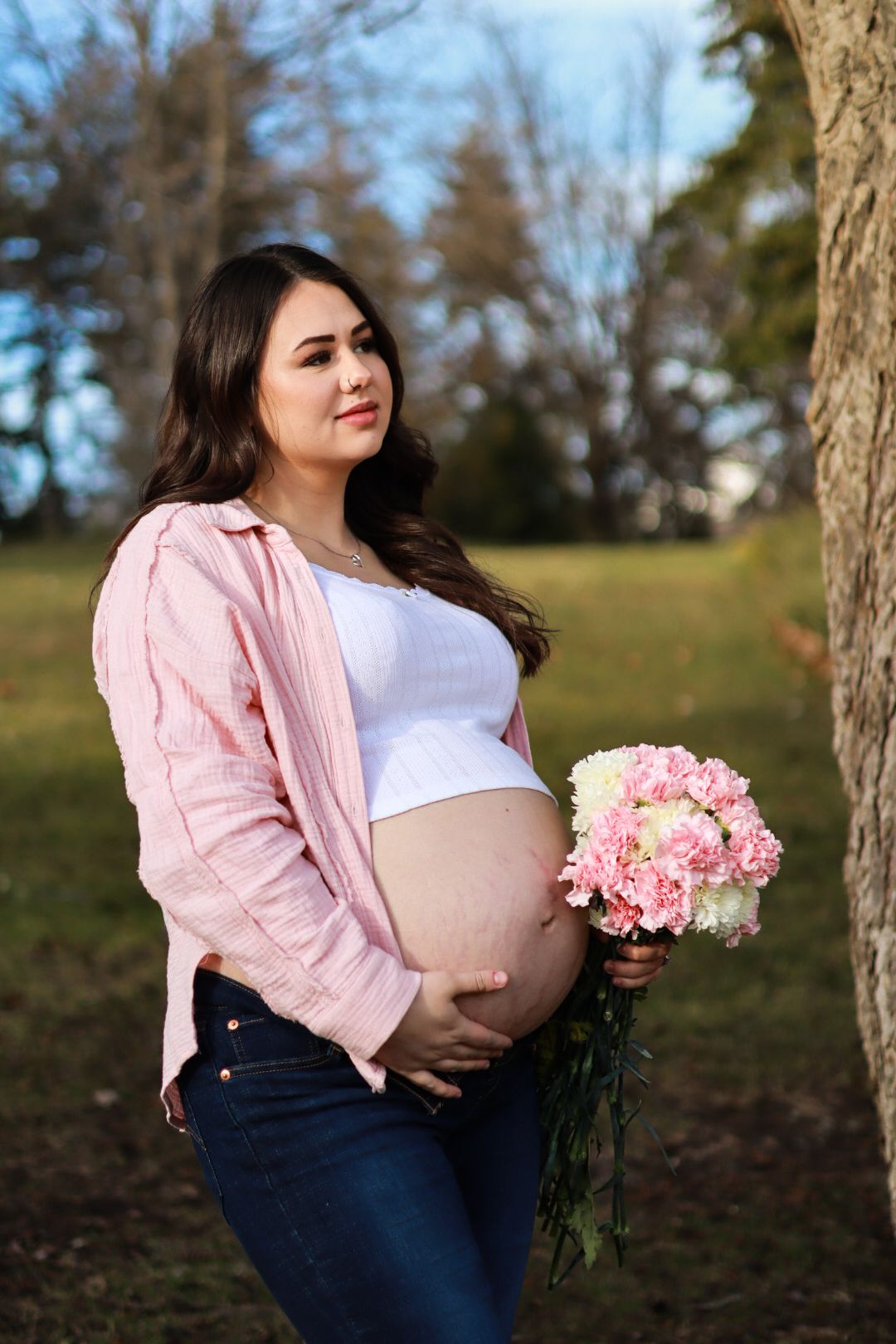 Expectant mother in pink cardigan and white top holds pink flowers while posing outdoors in a natural setting.