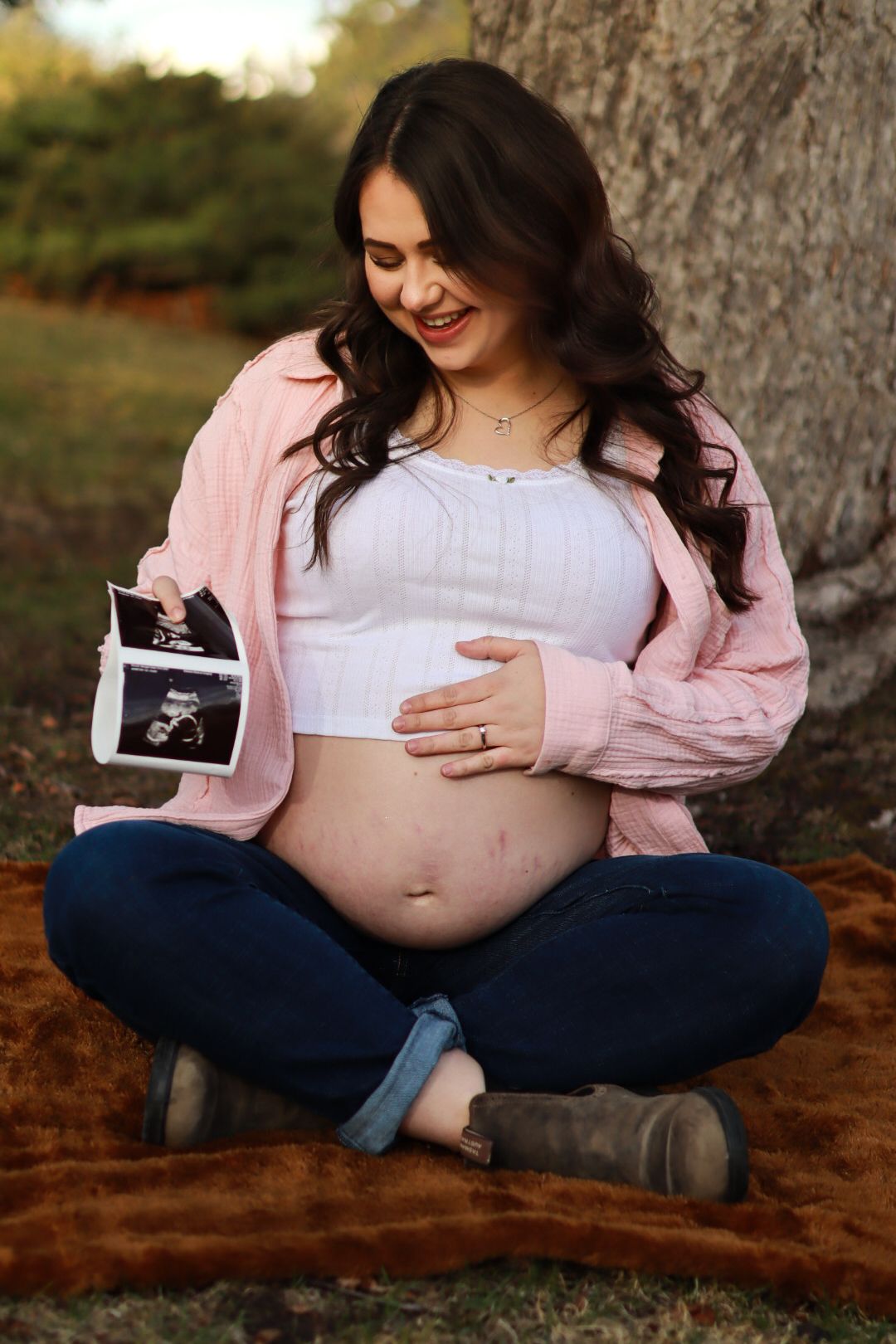 Maternity photo of a woman in pink jacket holds sonogram photo while sitting cross-legged under a tree.