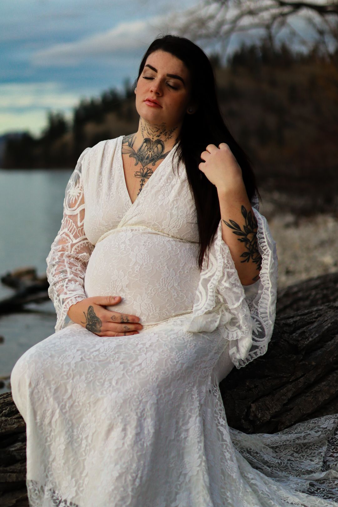 Maternity photo of woman in white lace dress sitting on rocks by a lake at sunset.