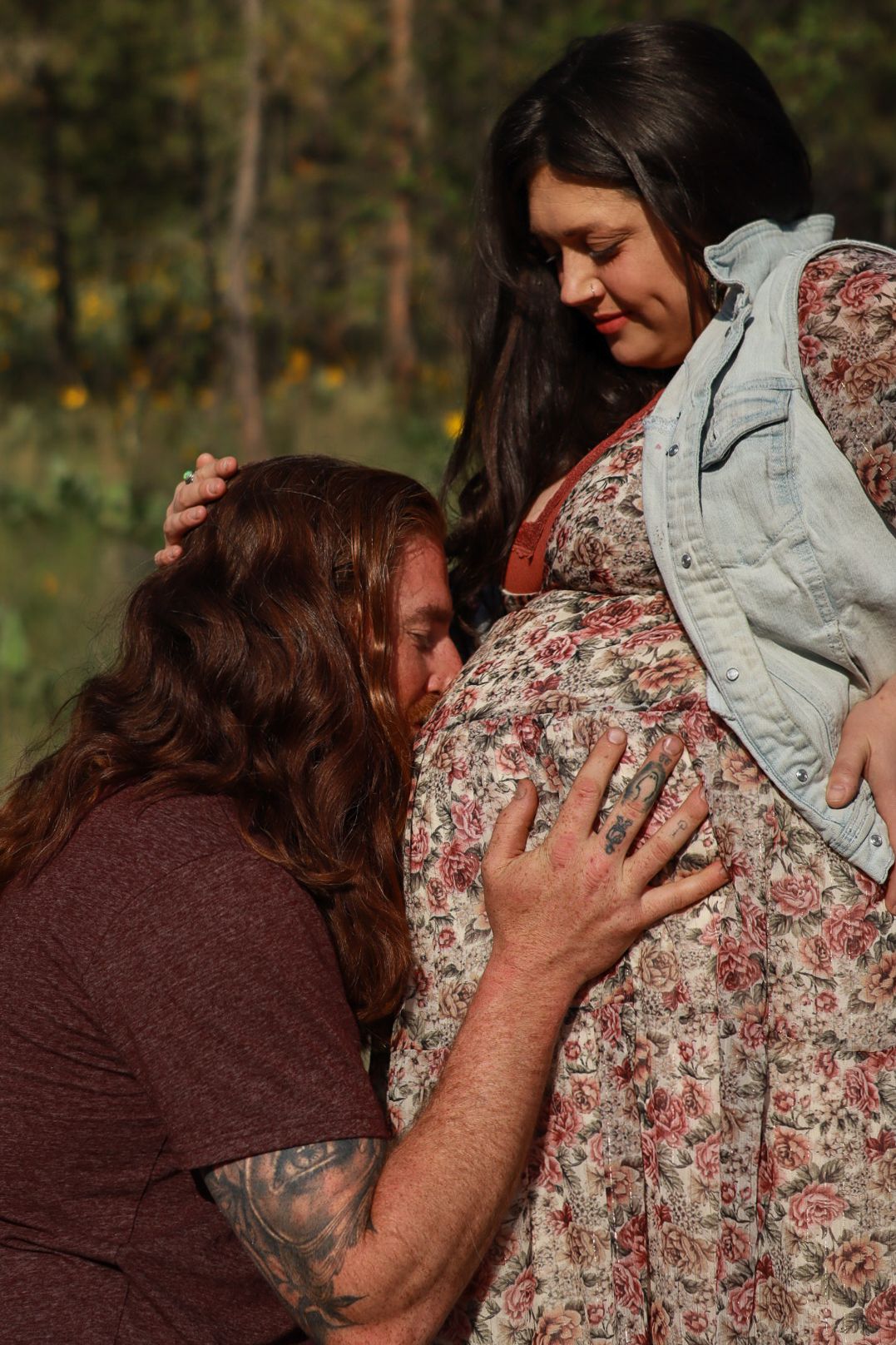 A tender pregnancy photo outdoors showing someone gently touching a pregnant belly in a floral dress and denim jacket.