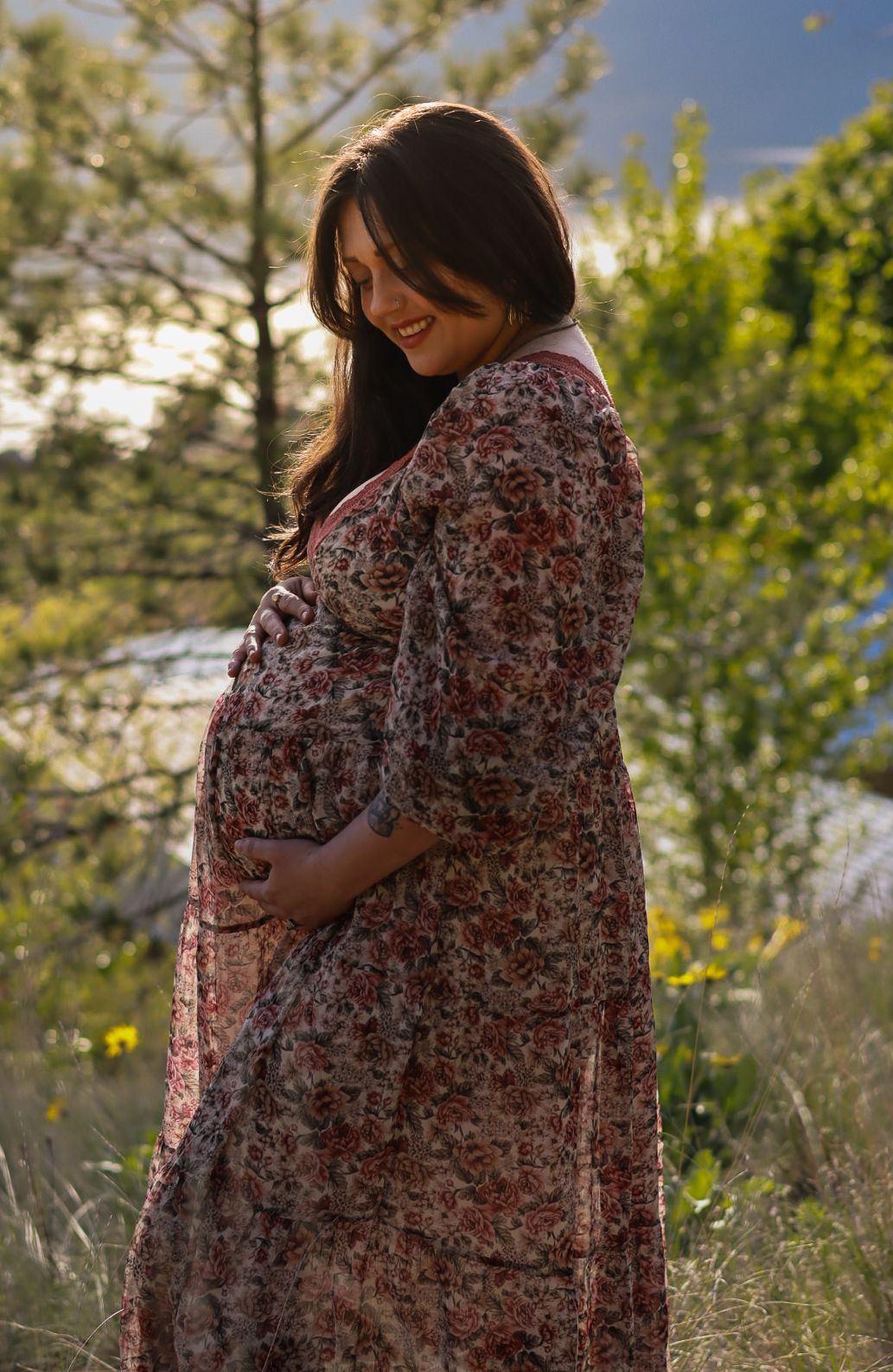 Pregnant woman in floral dress stands in sunlit meadow with wildflowers and trees in background.