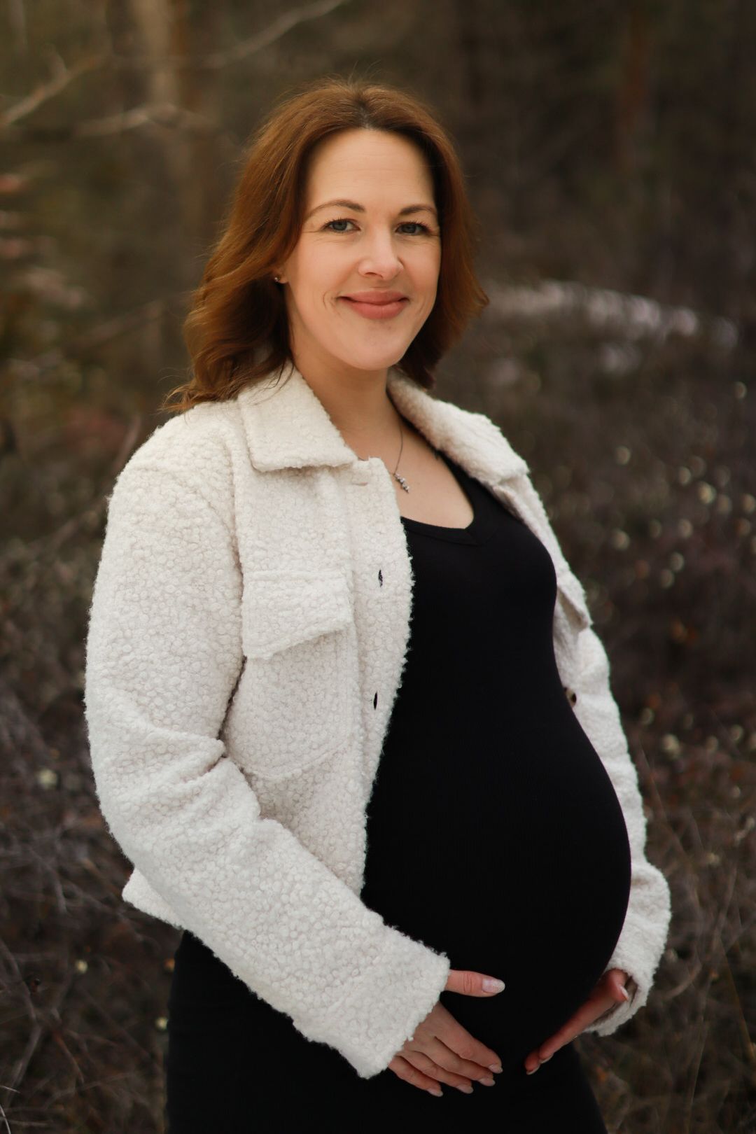 Person in cozy white teddy coat and black dress cradles pregnant belly while standing outdoors in nature.