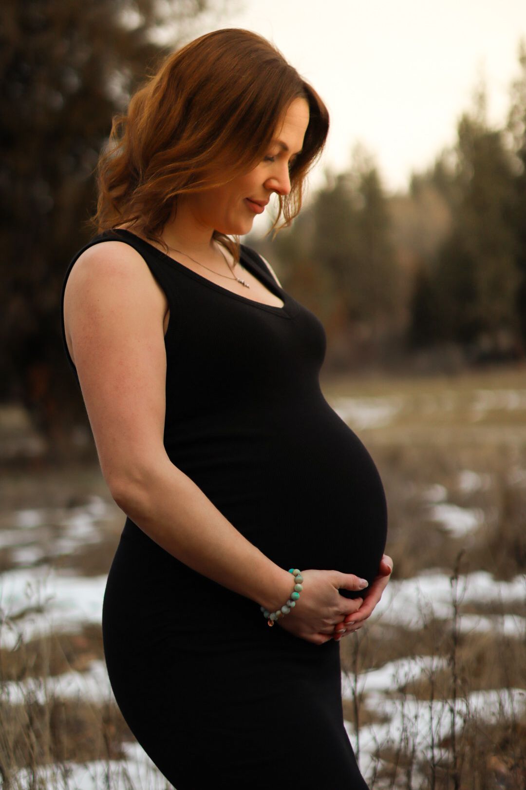 A pregnant woman in a black dress stands outdoors during winter, lovingly cradling her belly.