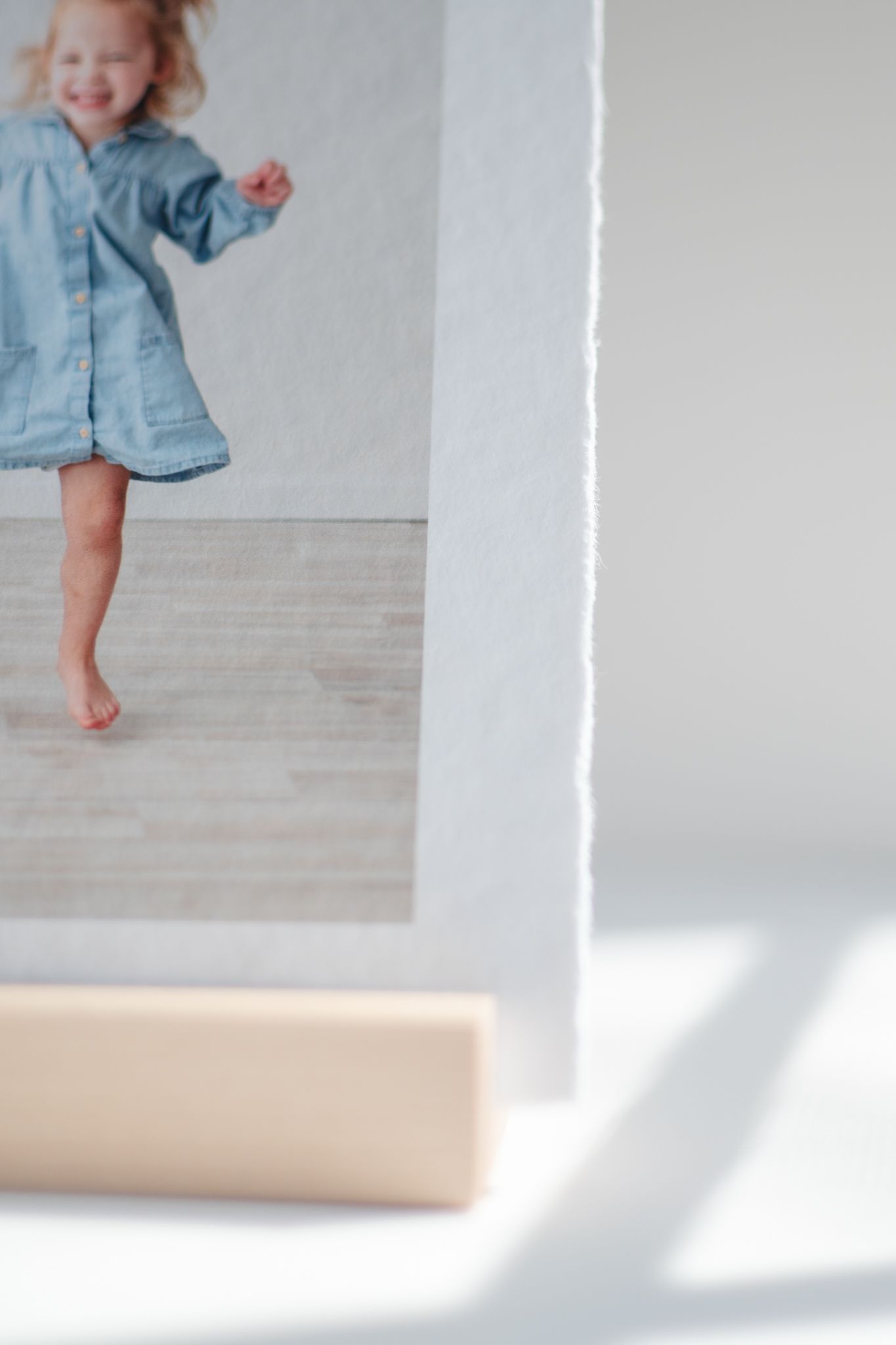 A young child in a denim dress twirls joyfully barefoot against a white wall with soft shadows.