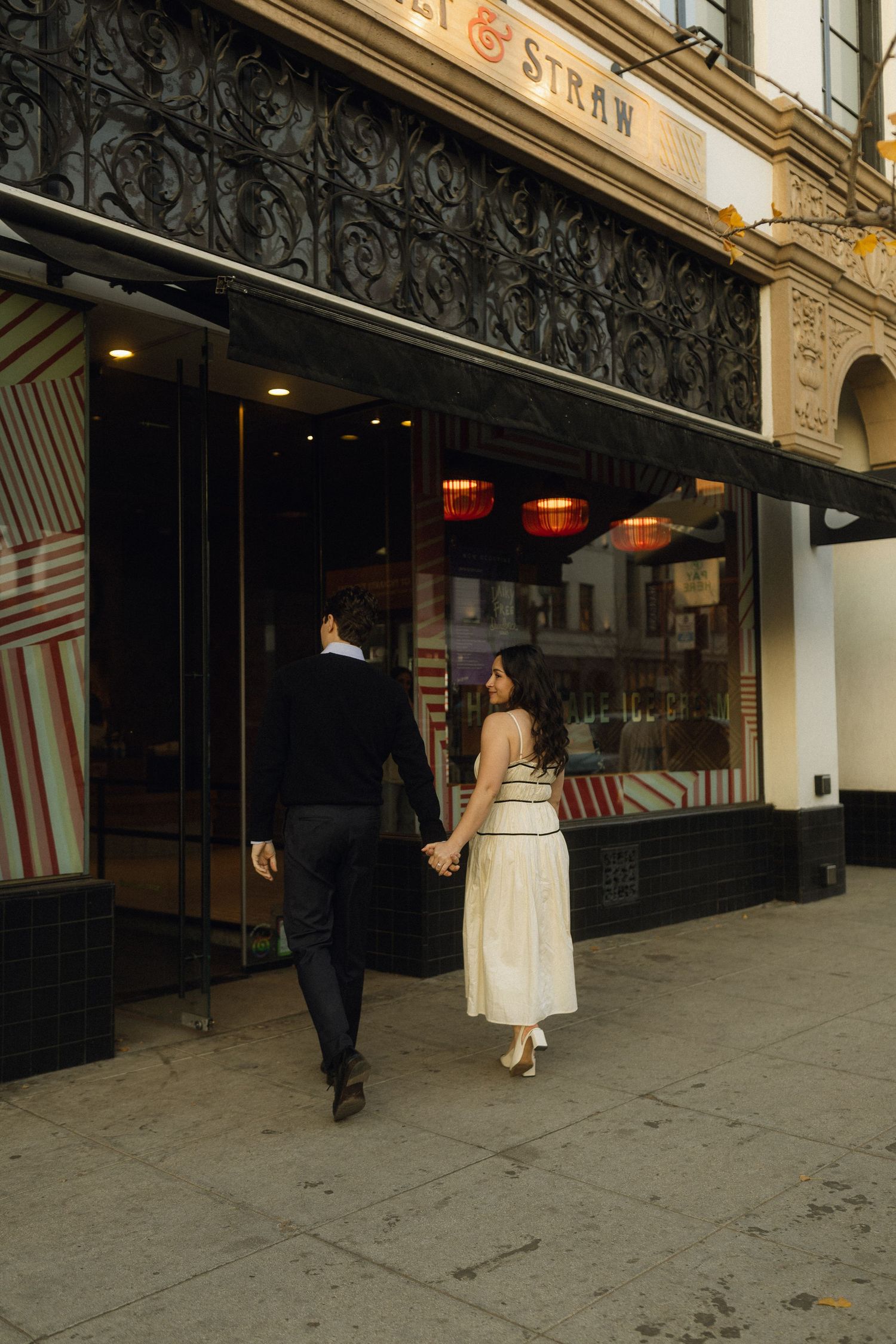 A couple walks together holding hands outside a vintage storefront with a decorative black facade and red lanterns.