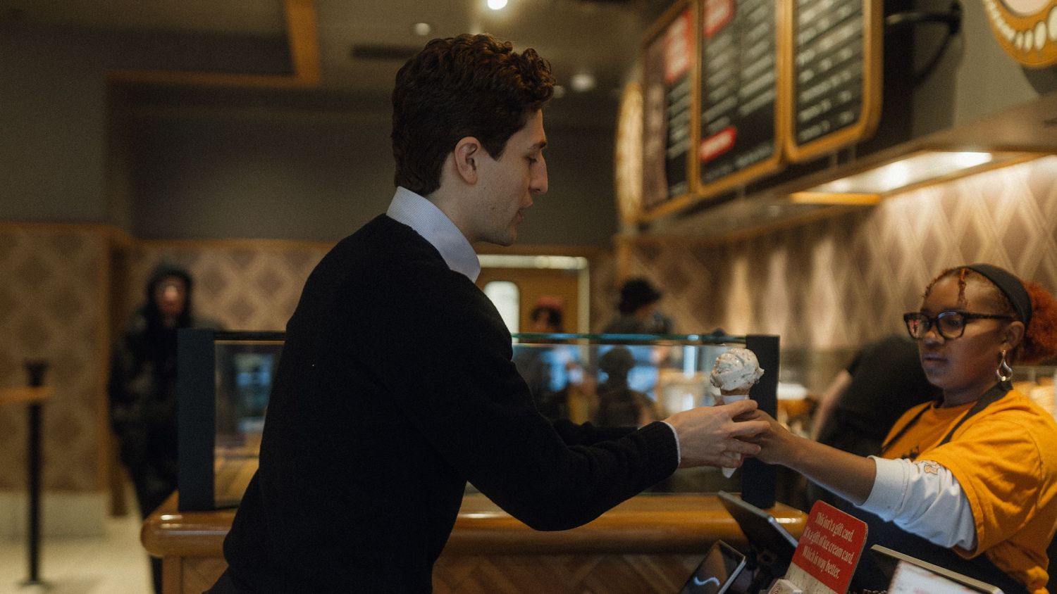A customer receives a coffee cup from a barista wearing an orange shirt at a coffee shop counter.