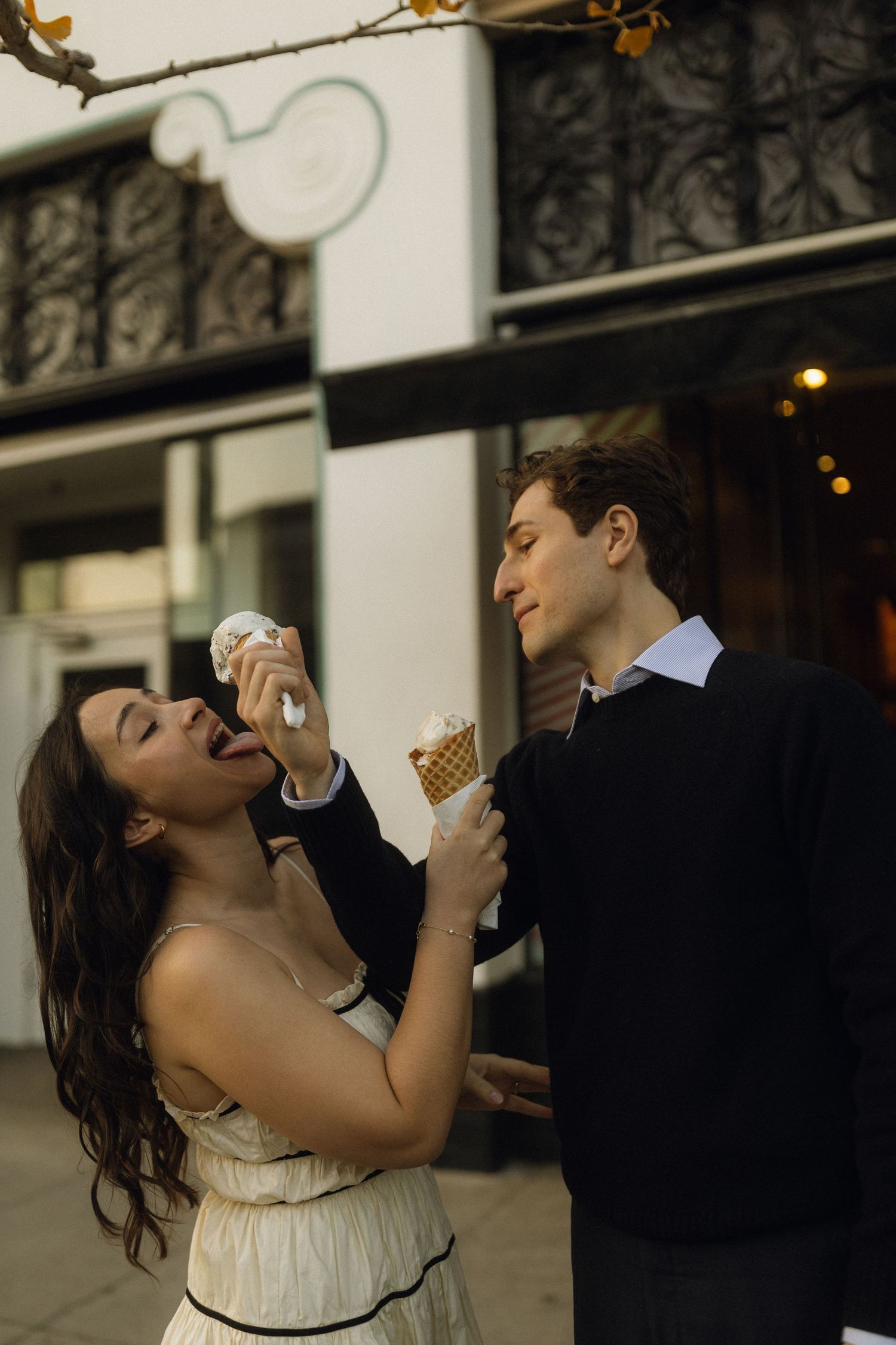 A couple shares ice cream cones and laughs together on a city street at dusk.