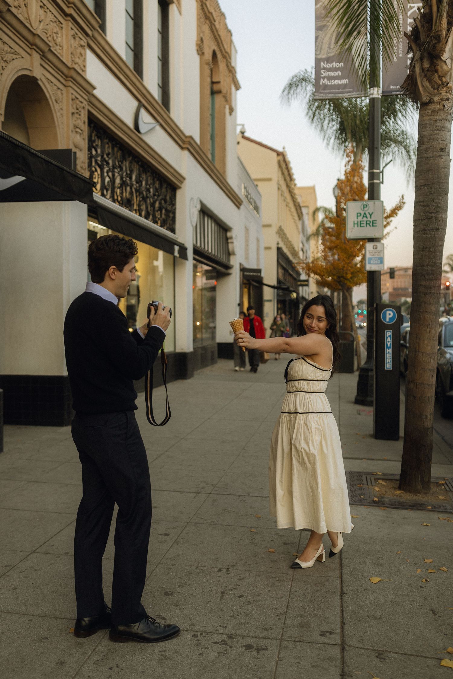 People in vintage-style clothing pose for photos on a palm tree-lined street with classic architecture.