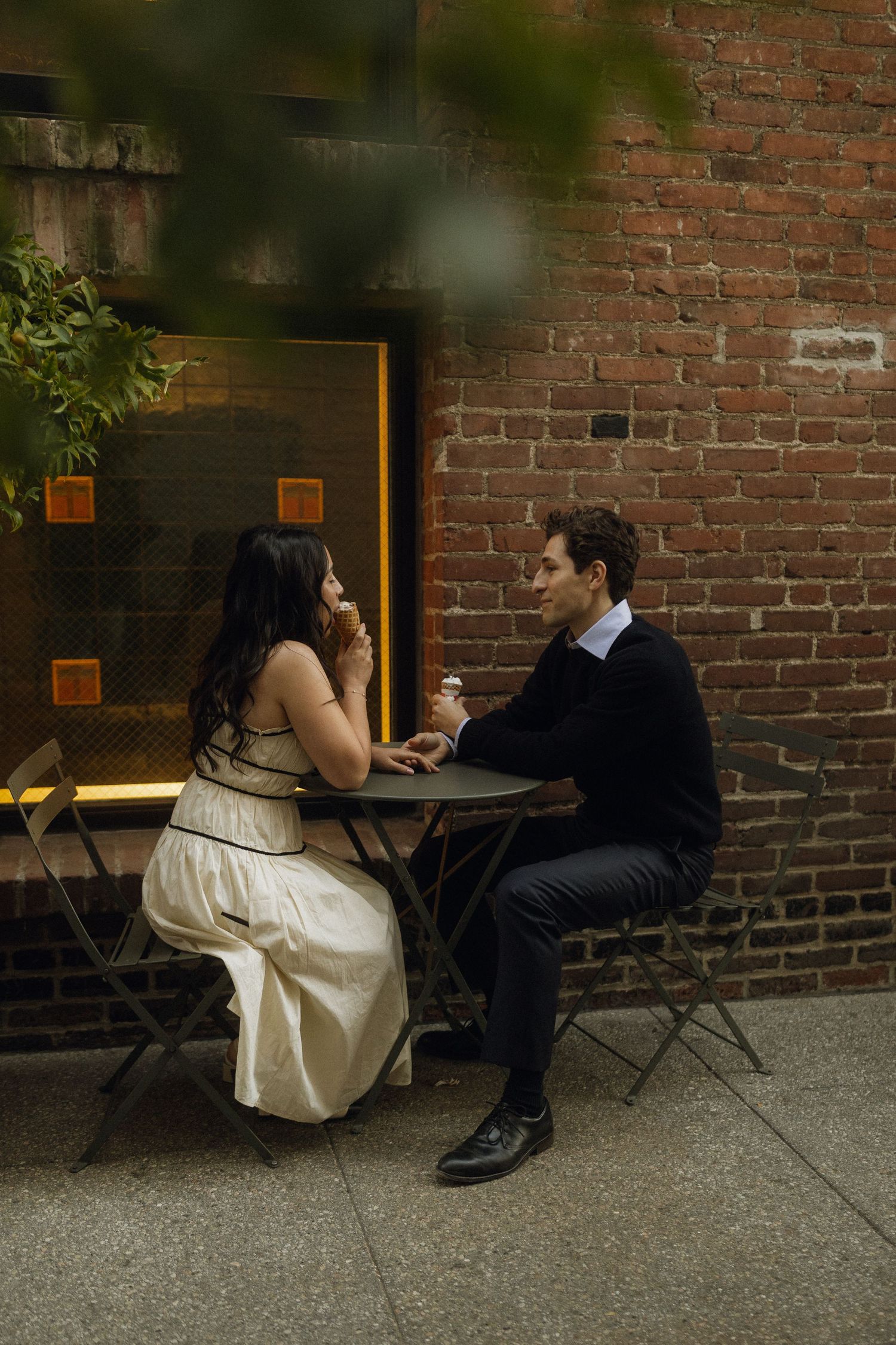 A couple sits intimately at a bistro table against a brick wall outside a restaurant on a warm evening.