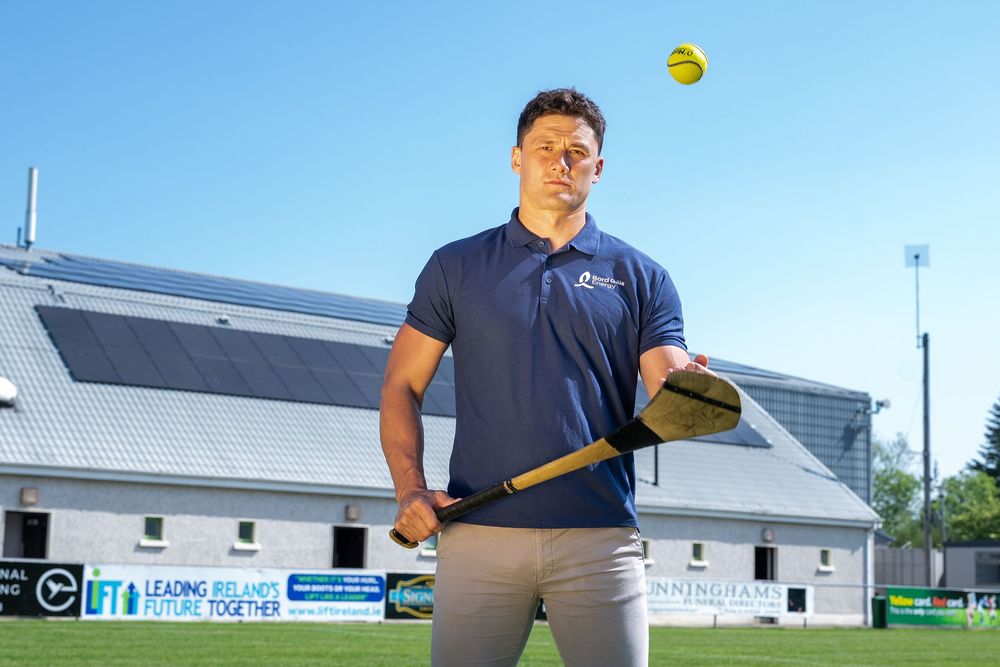 Man holding a hurling stick with a sliotar ball in the air at an outdoor GAA sports ground on a sunny day.